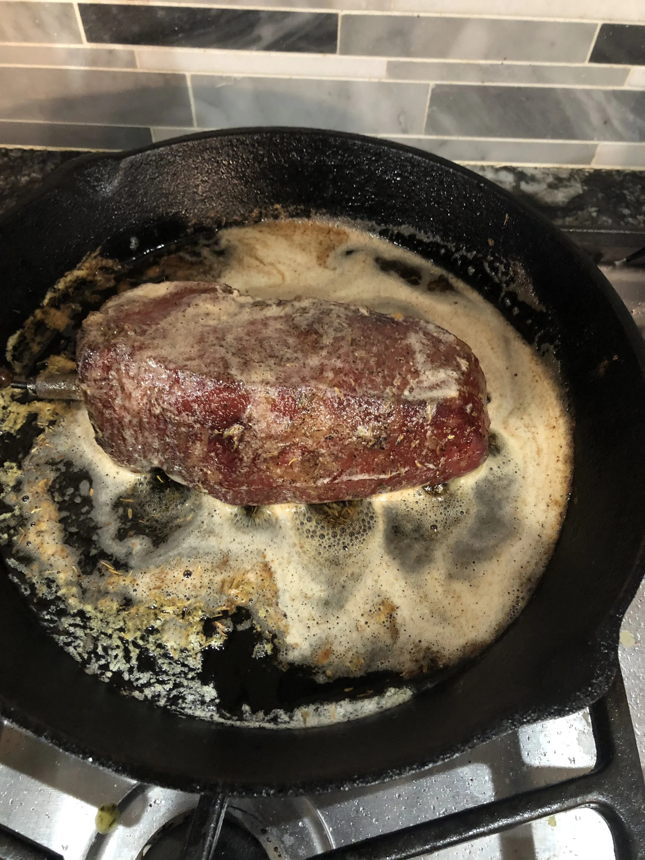 A cast iron skillet with a raw piece of beef cooking in a creamy sauce on a stovetop.
