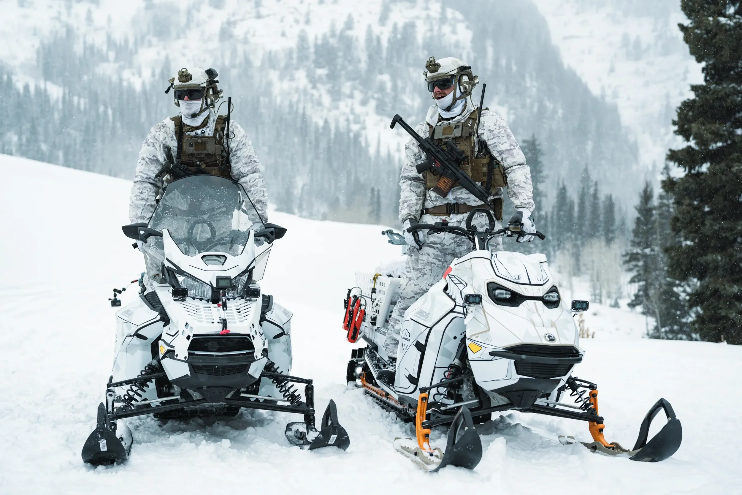 Two soldiers in camouflage uniforms riding snowmobiles in a snowy mountainous landscape.