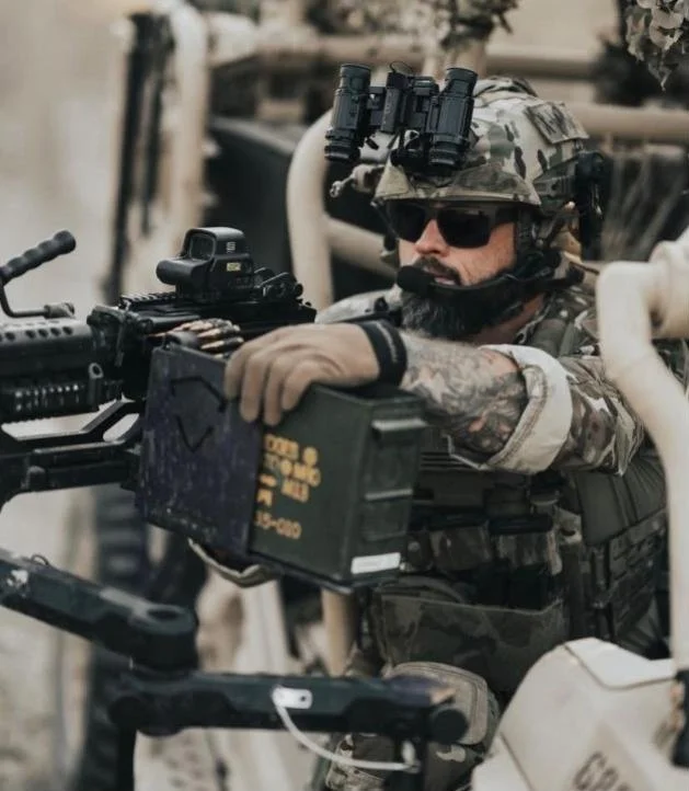 A soldier in tactical gear and sunglasses aiming a firearm with a mounted scope, wearing a camo helmet with night vision goggles, in a desert environment.