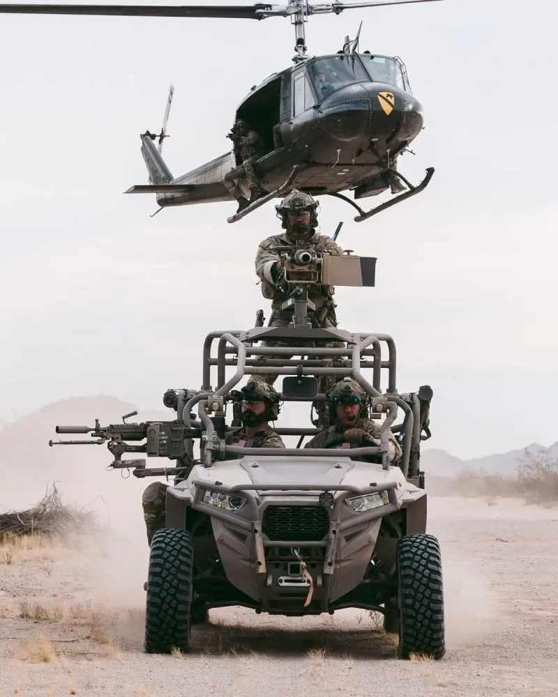 Military personnel riding an ATV with mounted weapons, a soldier operating equipment standing on top, and a black helicopter flying overhead in a desert landscape.