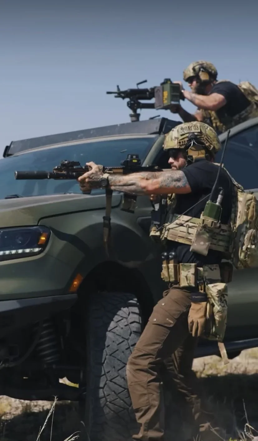 Two soldiers aiming rifles from behind a military vehicle during daytime.