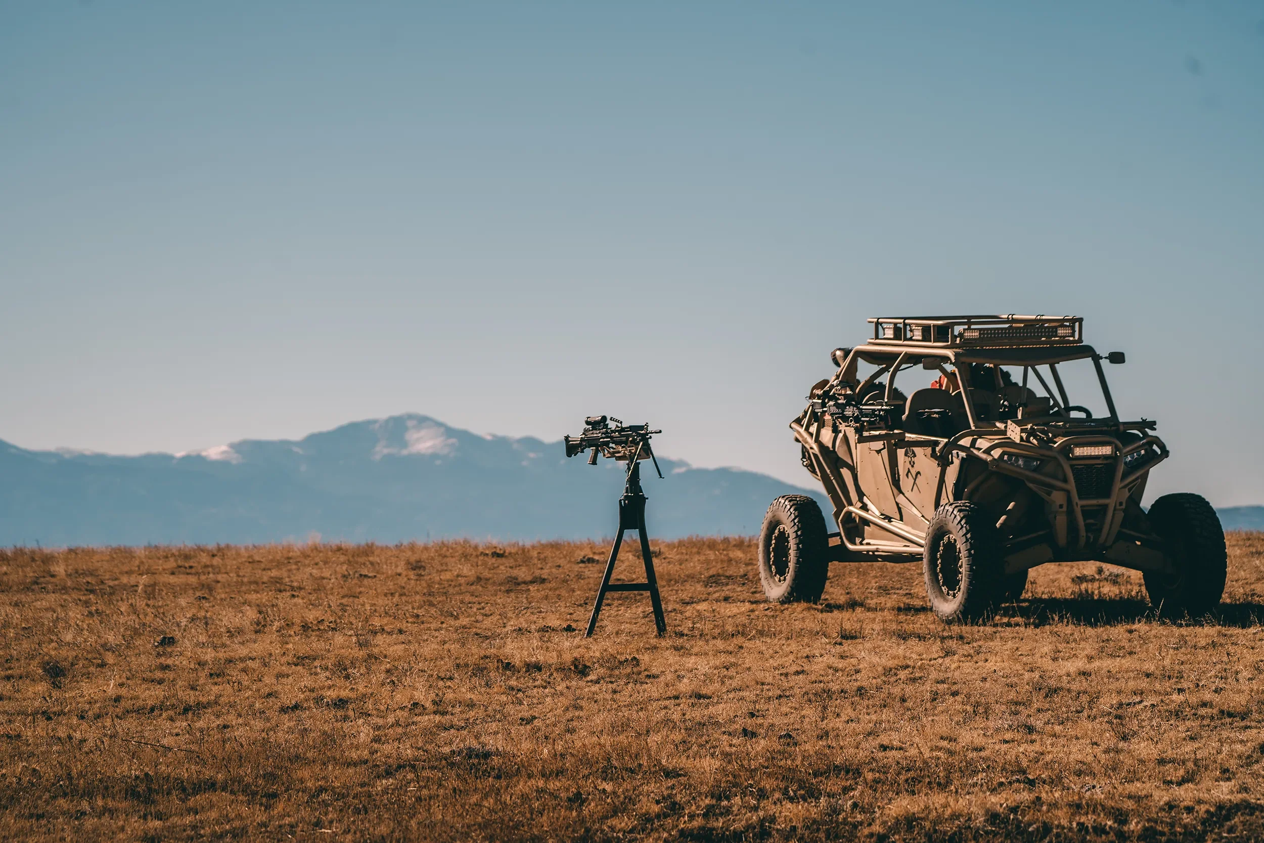 Off-road vehicle with mounted machine guns parked on a grassy plain with mountains in the background and a camera set up nearby, under a clear blue sky.