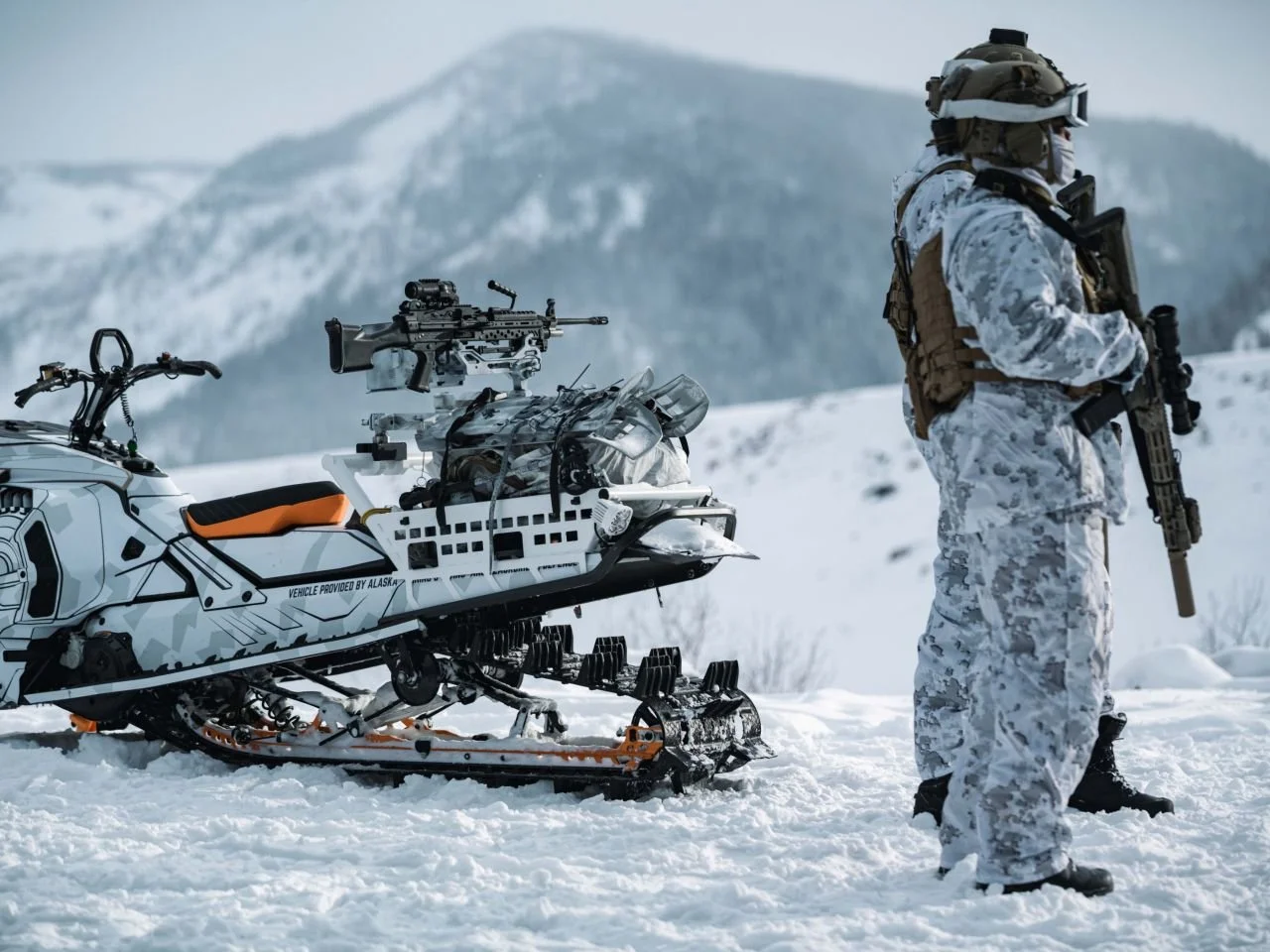 A soldier in camouflage winter gear standing in snowy landscape next to a snowmobile equipped with a mounted rifle.