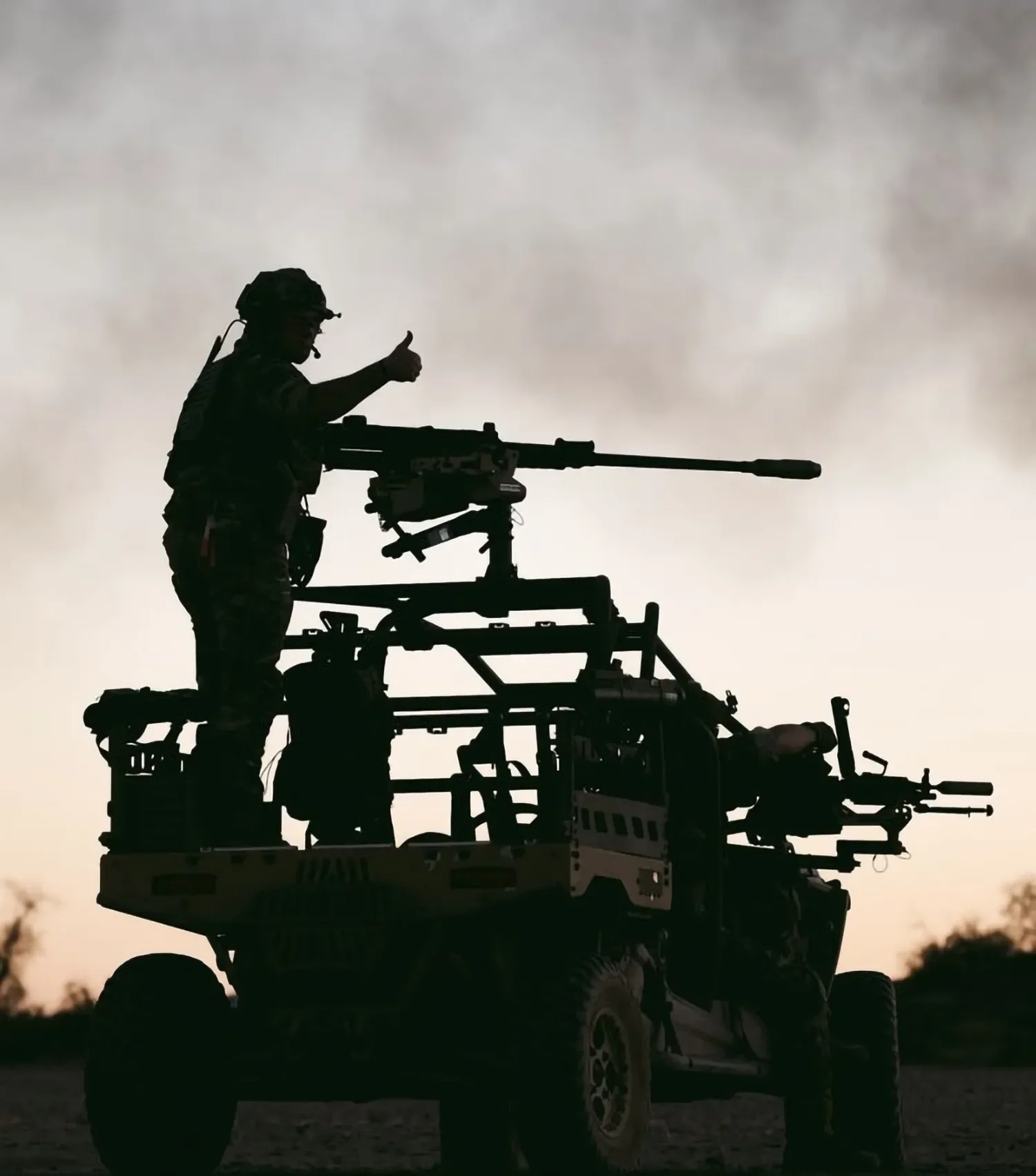 Silhouette of a soldier giving a thumbs up while operating a mounted machine gun on a military vehicle during sunset.