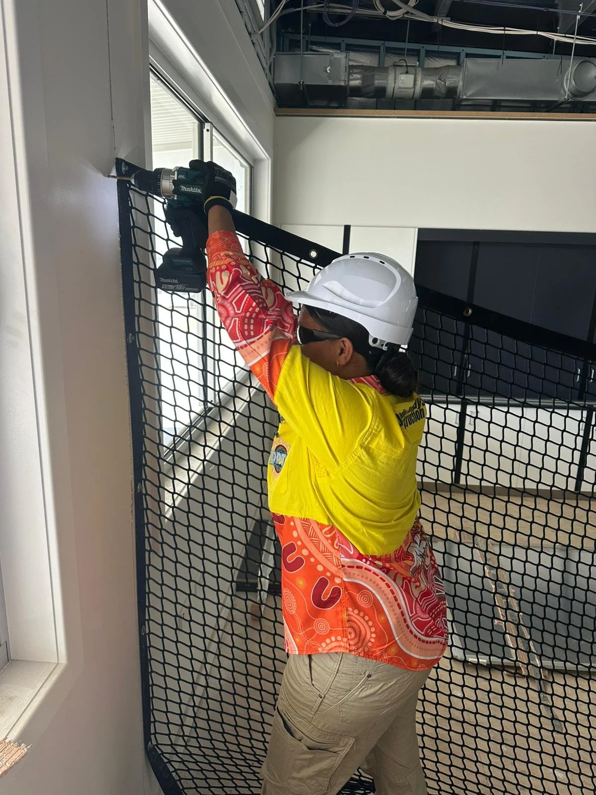 A person wearing a white safety helmet, sunglasses, a yellow safety vest, and a colorful patterned shirt is fixing a metal mesh partition to the wall with a power drill inside a room with exposed ductwork.
