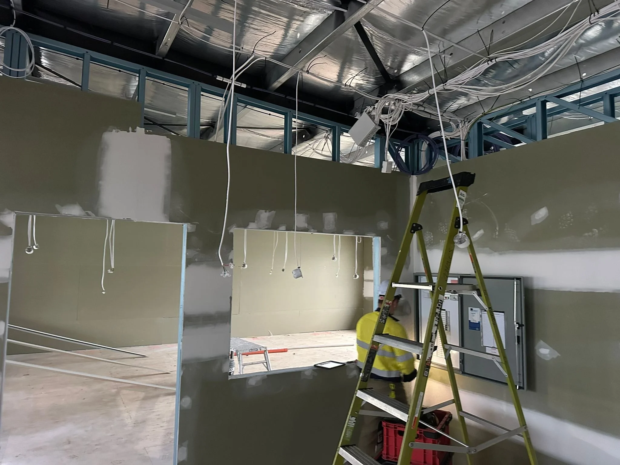 Construction site inside a building with drywall, a ladder, and exposed ceiling with wires and ductwork.