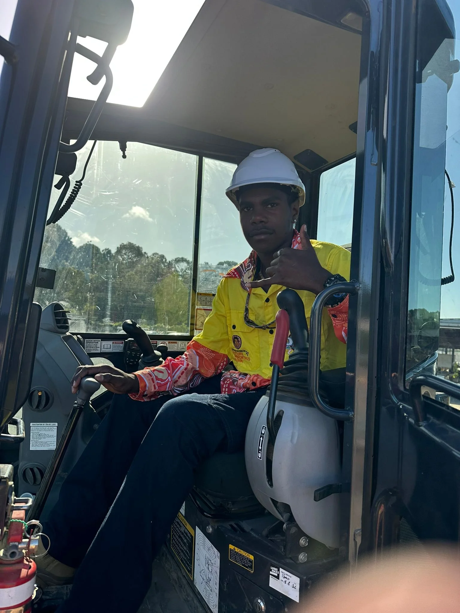 Man wearing a yellow safety vest and a white safety helmet, sitting inside a construction vehicle, making a thumbs-up gesture.