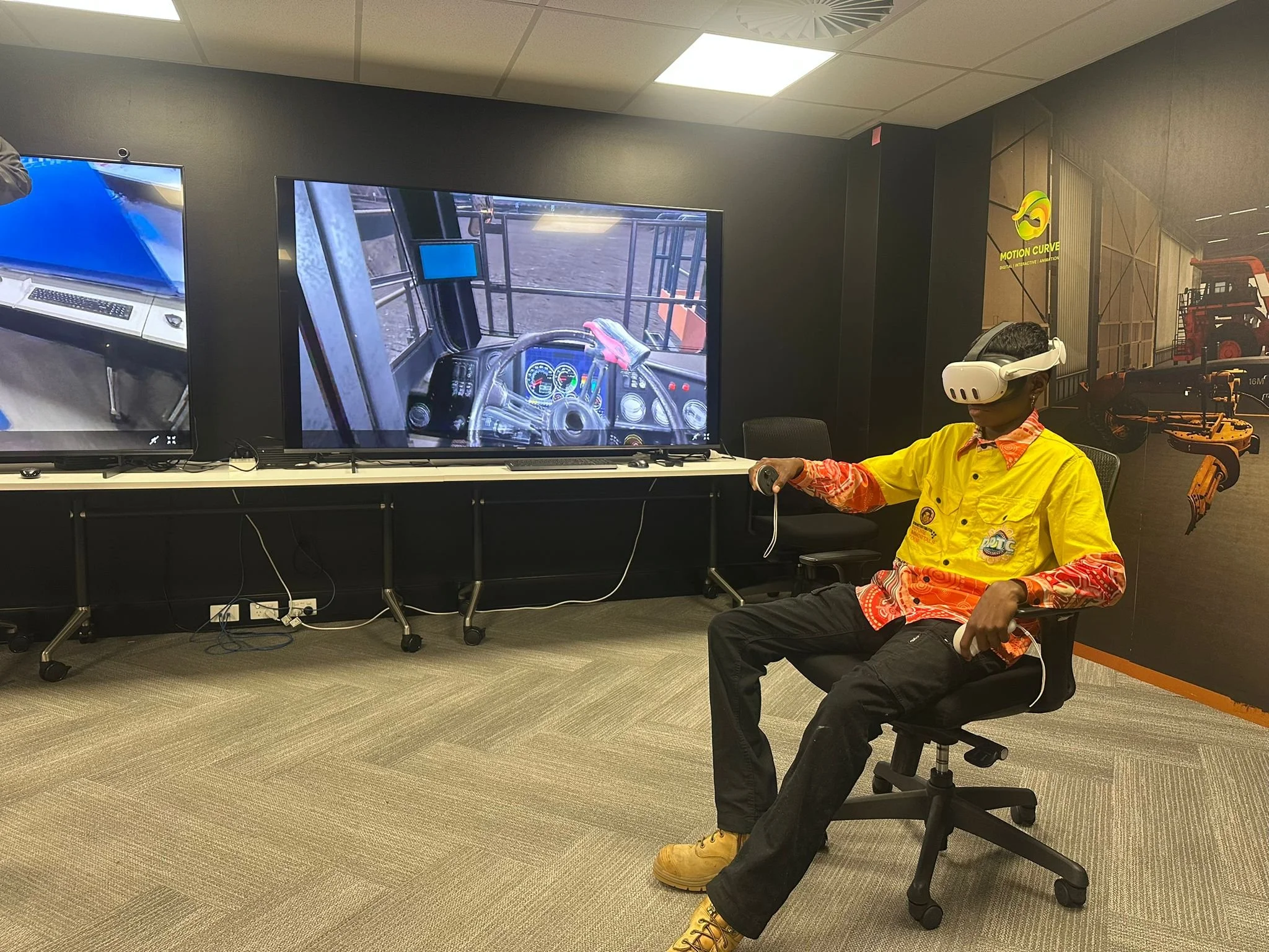 A person wearing a yellow jacket and a virtual reality headset, sitting on a chair in a room with large screens displaying a driving simulation. There are equipment and machinery in the background.