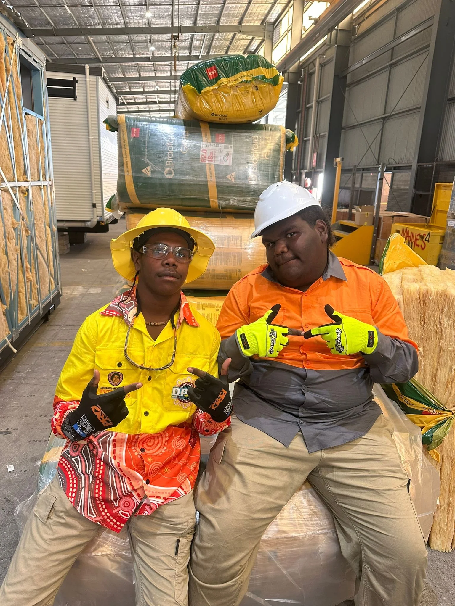 Two workers wearing safety helmets and gloves in a warehouse or industrial setting, posing with their hands pointing towards each other. They are dressed in colorful shirts, with one in yellow and the other in orange, standing in front of large wrapped packages and industrial equipment.