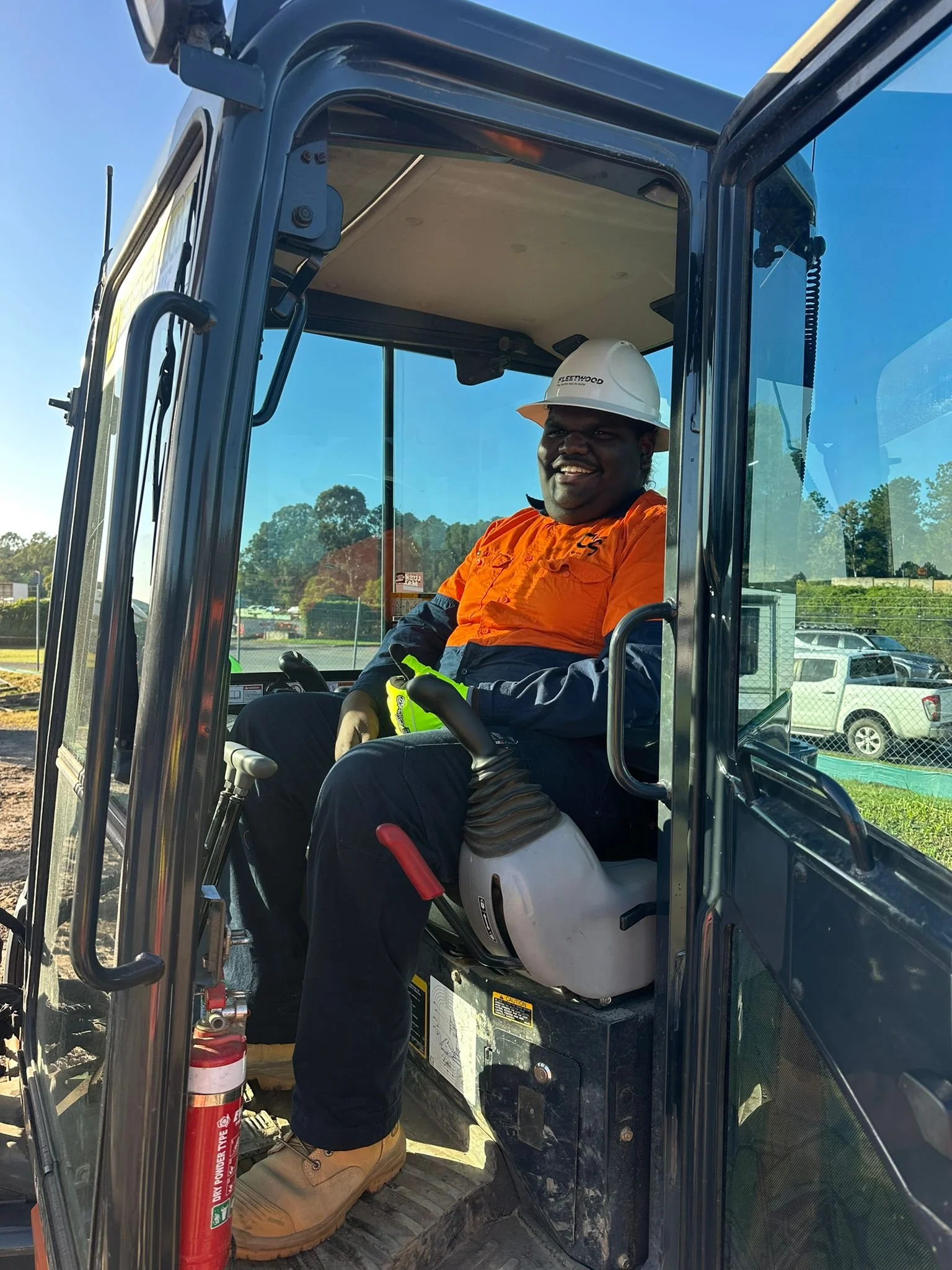 A man sitting inside a construction vehicle, wearing a white helmet and orange high-visibility shirt, smiling at the camera.
