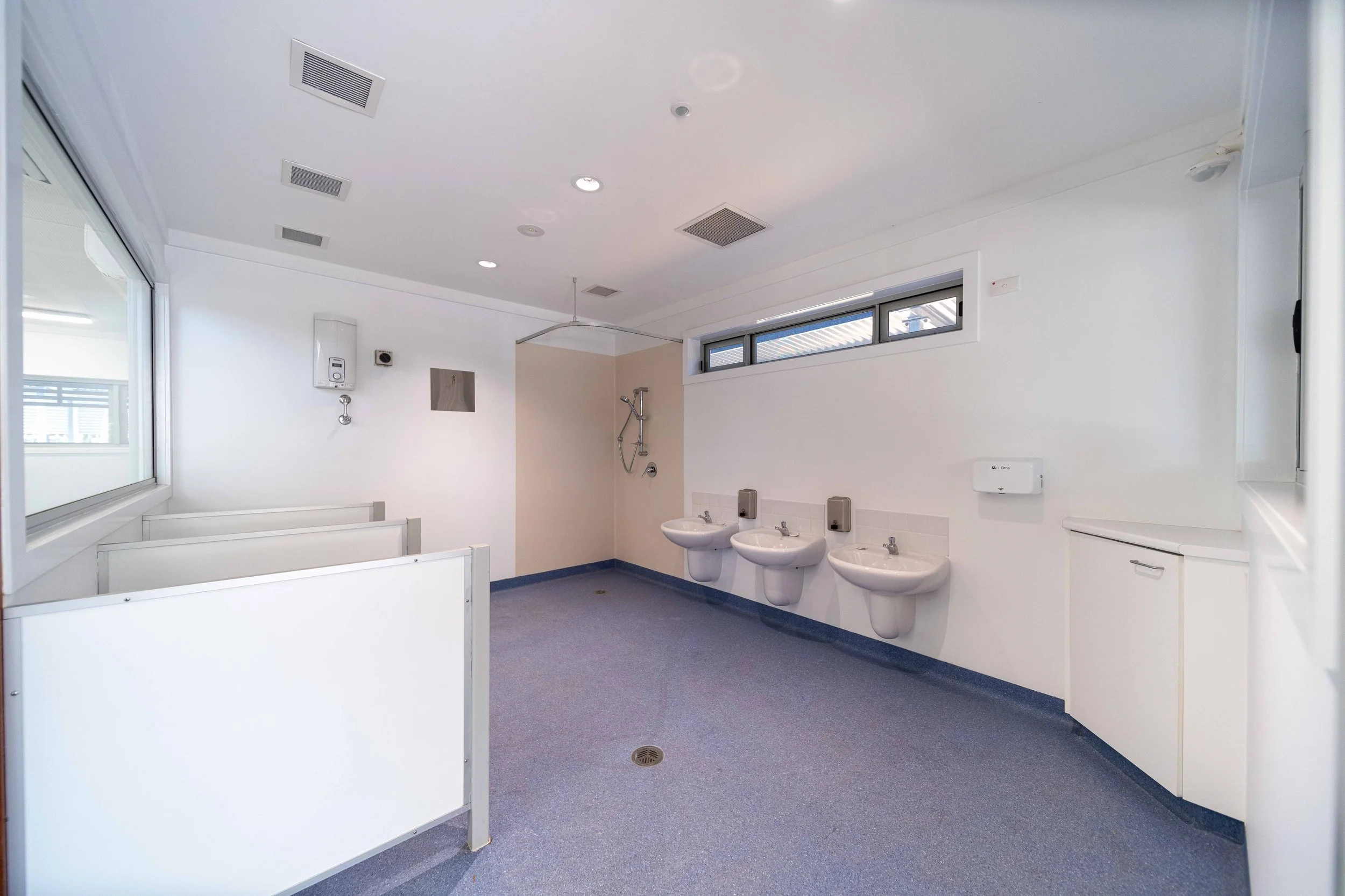 Public restroom with three sinks, hand dryers, and a shower area, with a window and ceiling vents.