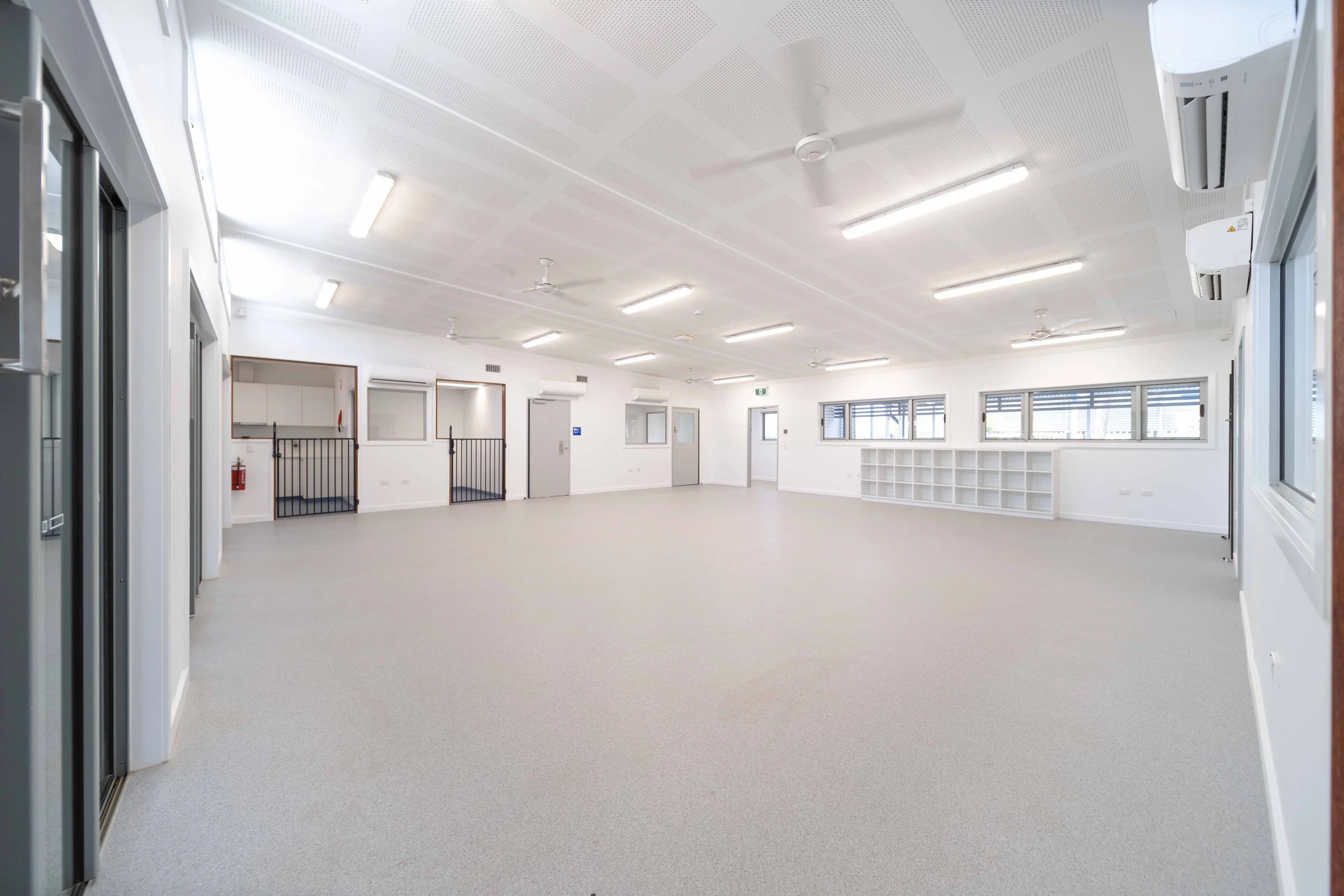 Empty classroom or community space with white walls, ceiling fans, large windows, built-in shelves, and bright overhead lighting.