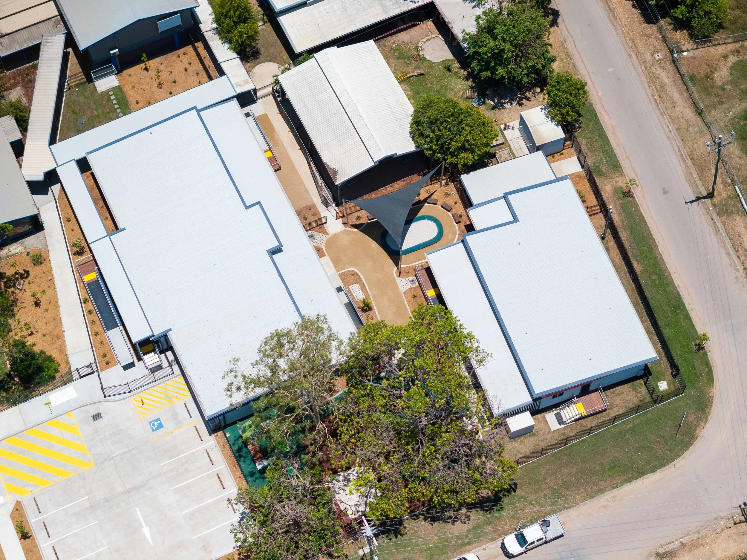 Bird's eye view of a school or community center with several white-roofed buildings, a parking lot with yellow-striped spaces, a playground with trees, a small pool, and surrounding roads.