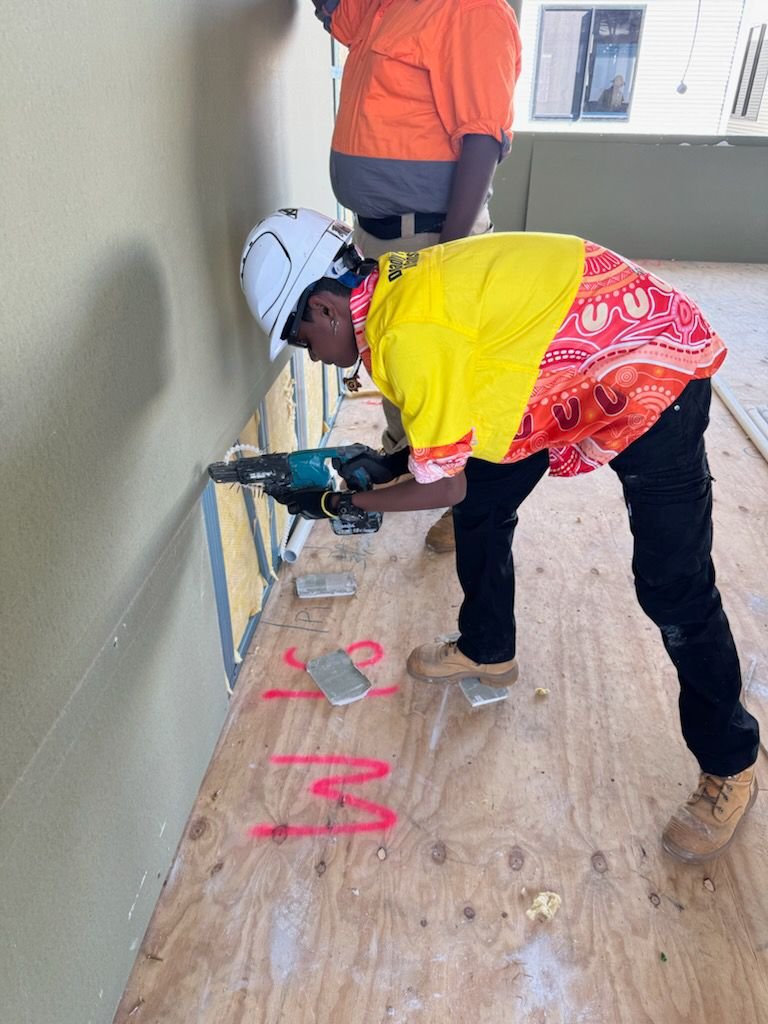 Construction worker wearing a yellow safety vest, patterned shirt, and white helmet using a power drill on a wall at a construction site.