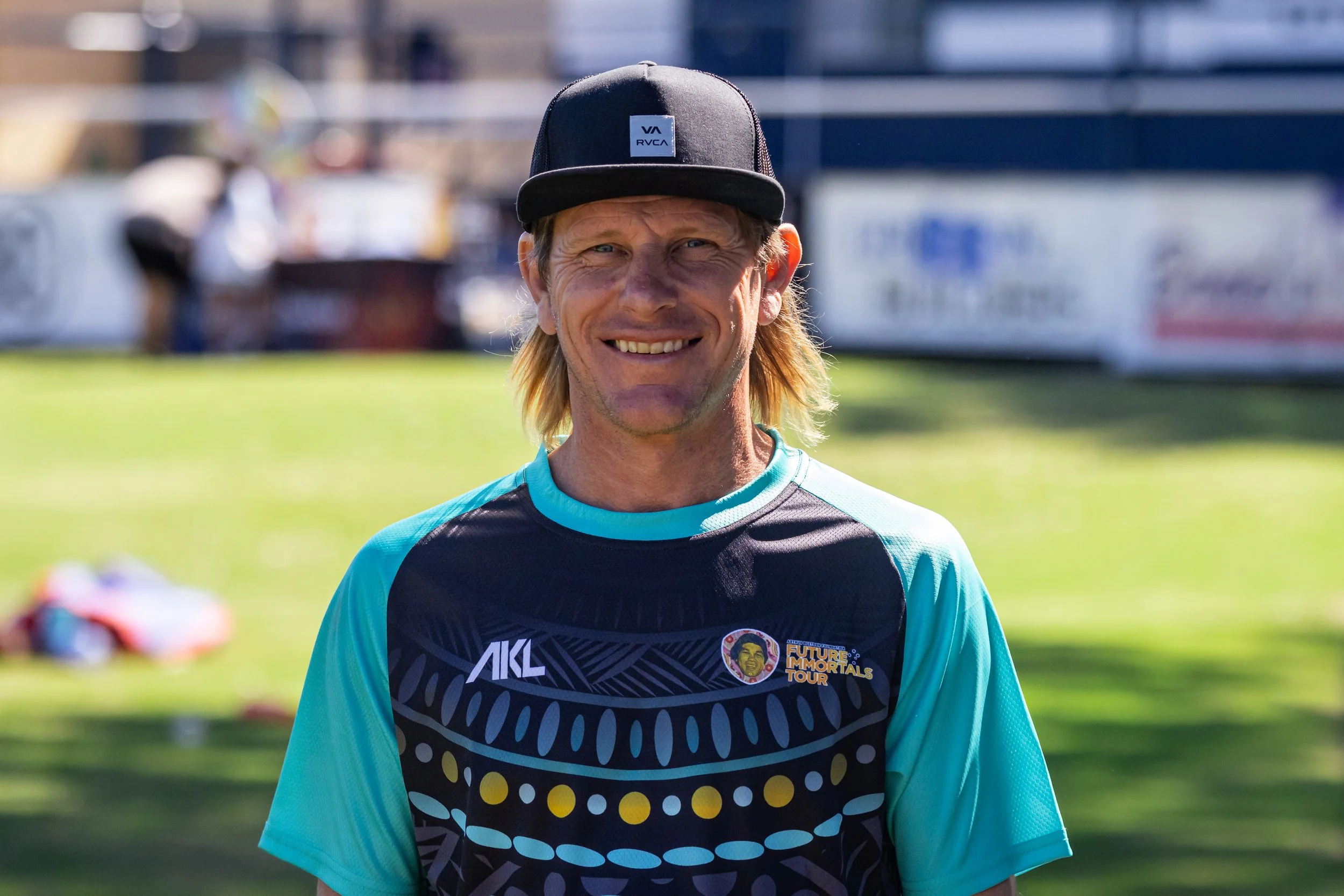 Smiling man wearing a black trucker hat and athletic shirt, standing outdoors on a sunny day with a blurred background of a football field and vehicles.