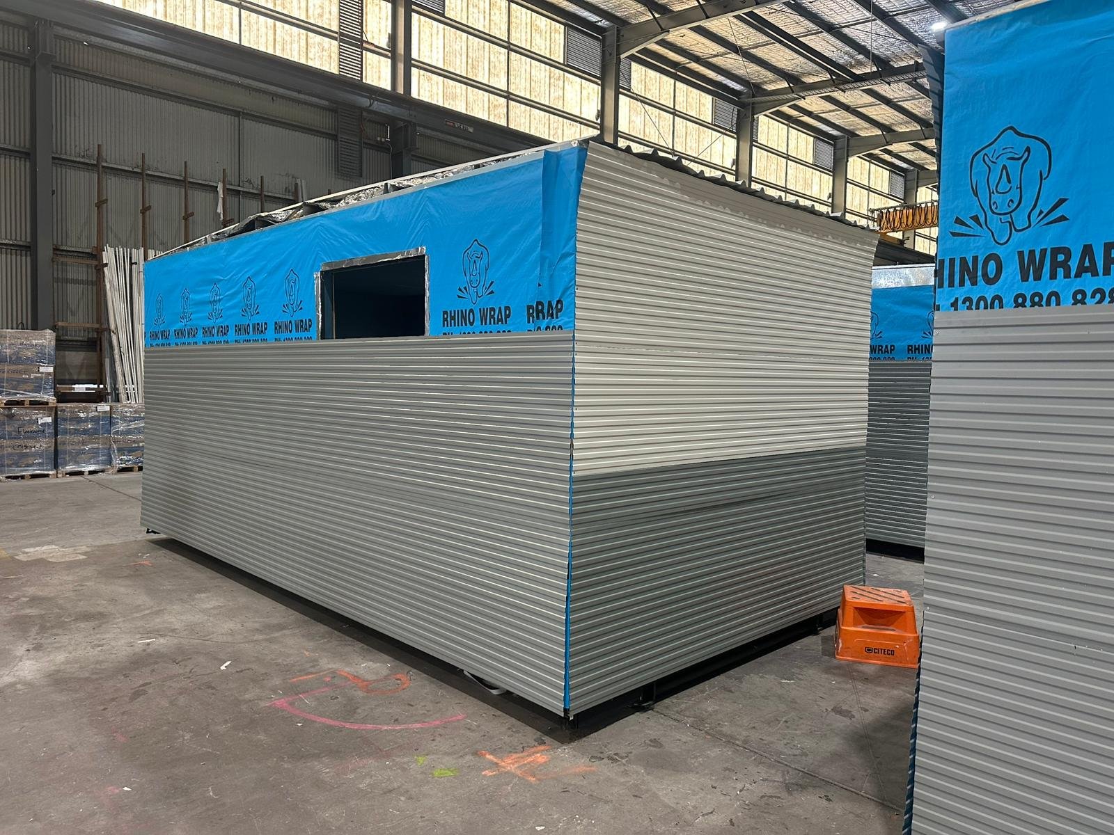 View of a construction site with a partially built small structure covered with blue Rhino Wrap and silver corrugated metal panels. The structure has an opening for a window, and there are construction materials and equipment nearby inside a warehouse with a high ceiling and metal framing.