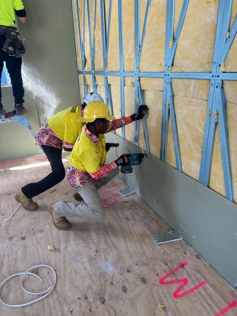 Two construction workers wearing yellow safety vests and helmets are installing metal flashing along a drywall wall inside a building under construction. One worker is kneeling with a power tool, while the other is securing the structure