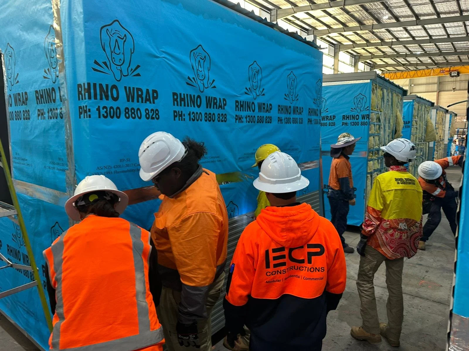 Group of construction workers in safety gear inspecting a large blue wrapped construction panel inside a warehouse.