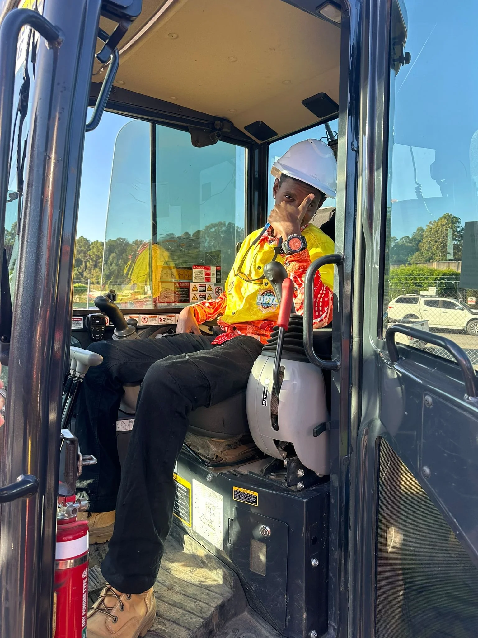 A person sitting in the driver’s seat of a small construction vehicle, wearing a white hard hat, yellow safety vest, and tan work boots, on a sunny day with trees and parked cars in the background.