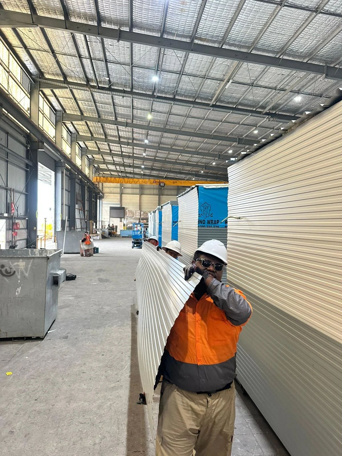 Workers in safety gear, including helmets and glasses, inside a large industrial warehouse, carrying long metal panels.