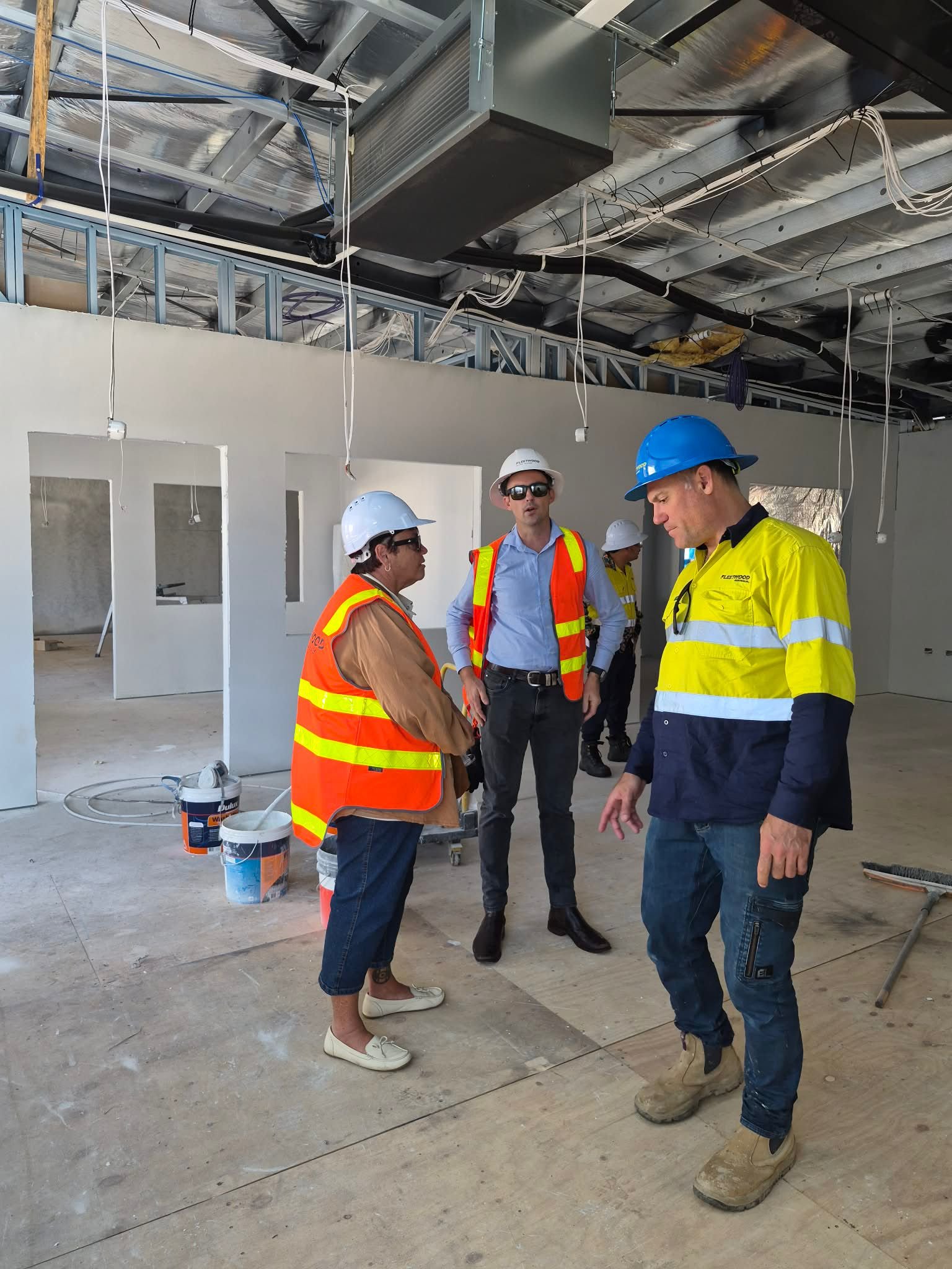 Construction workers wearing safety helmets and vests are inside a building under construction. They are standing on a concrete floor, with drywall, scaffolding, and construction materials visible around them.