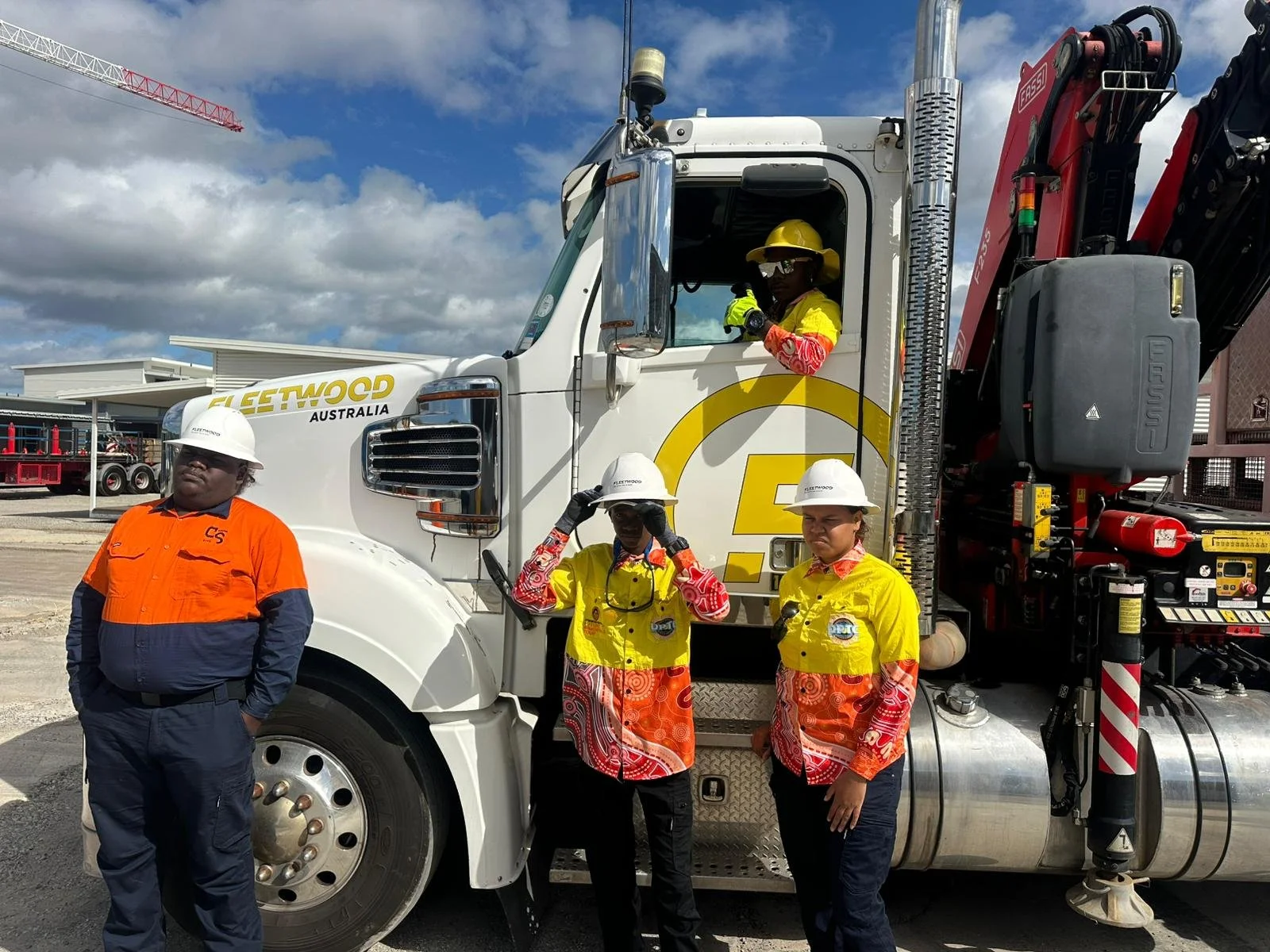 Four construction workers standing and sitting in front of a large white truck with a red crane in the background. Three workers are wearing yellow and orange safety uniforms with white helmets, and one is in an orange and navy uniform with a white helmet. The worker in the truck's window is waving.