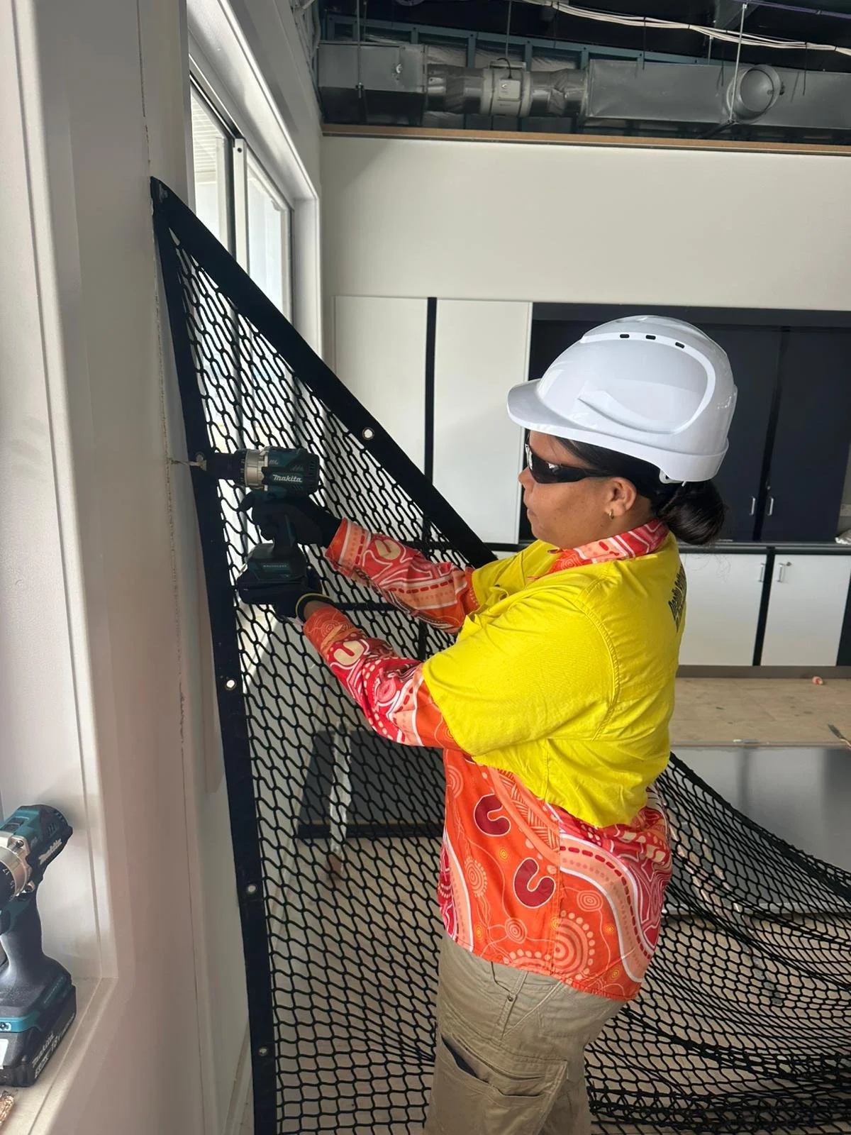 A woman wearing a white safety helmet, sunglasses, a bright yellow work shirt, and orange patterned cloth, using a power drill to work on a metal panel inside a construction site or industrial setting.
