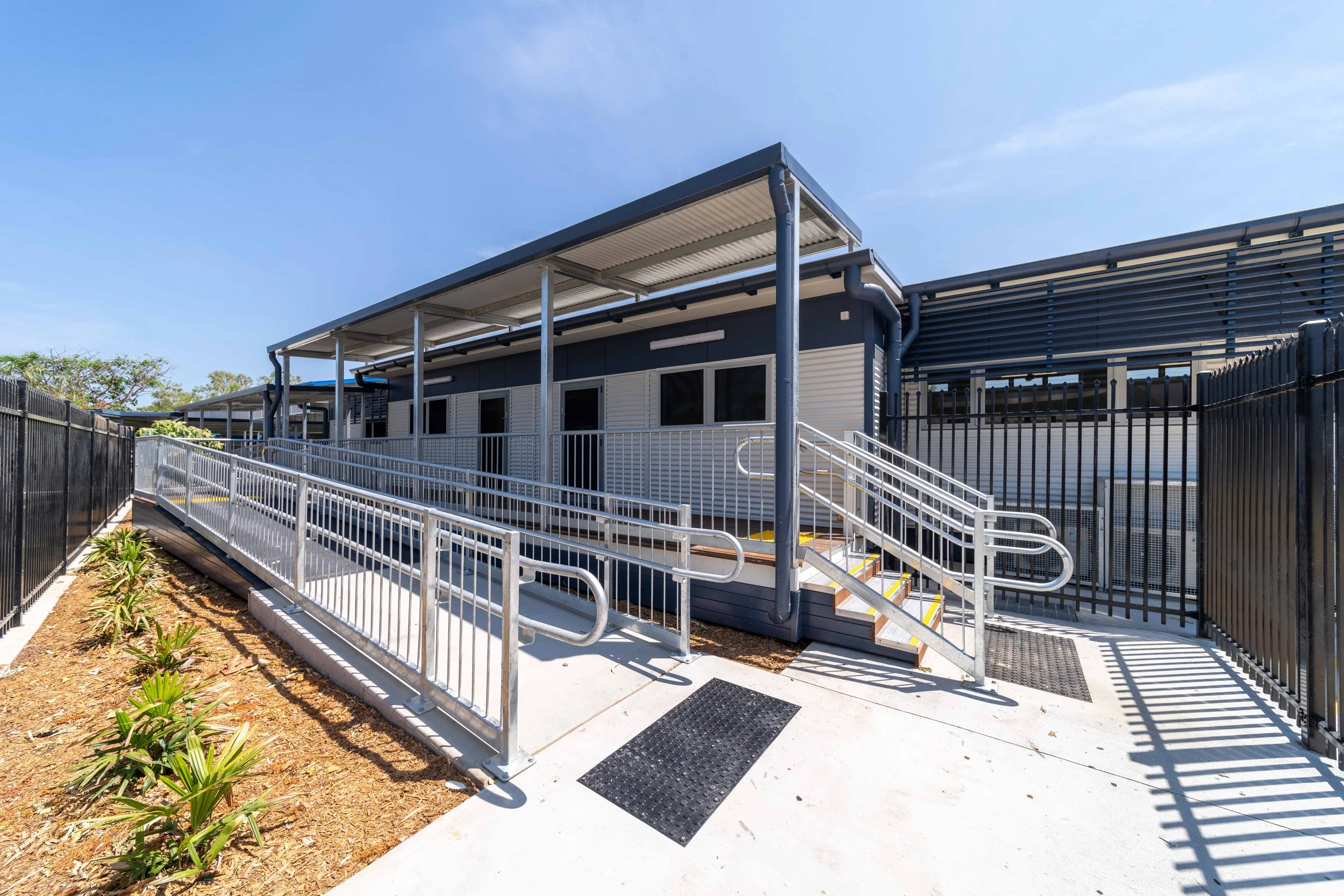 Accessible ramp leading to a modern building with metal railings, black fencing, and a blue sky.