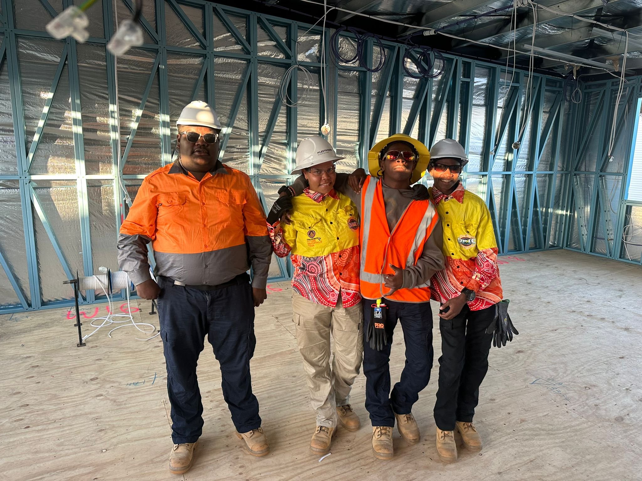 Five construction workers standing inside a building under construction with metal framing and exposed wiring, wearing safety gear including helmets and reflective clothing.