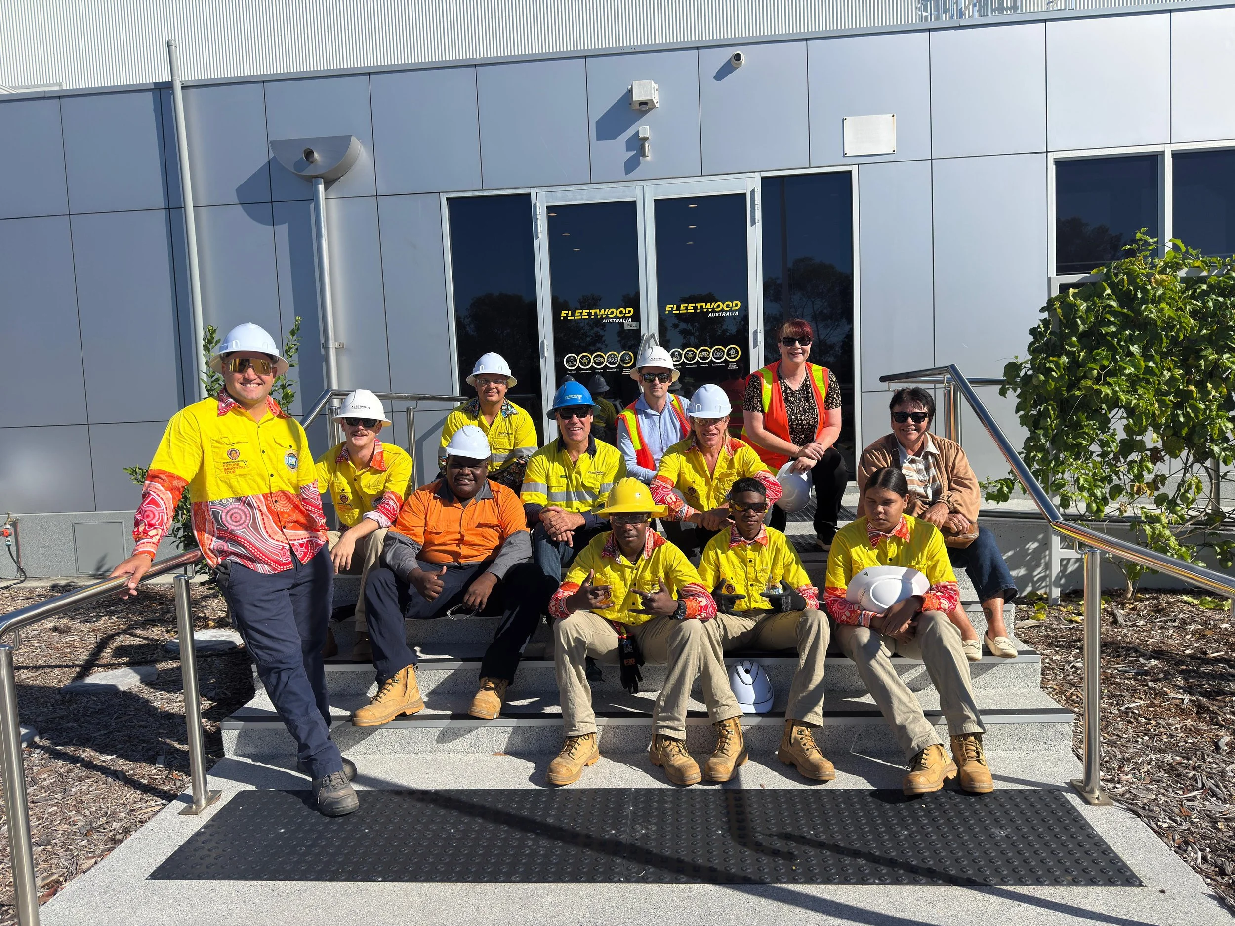 Group of workers, some wearing yellow safety shirts and white helmets, sitting and standing on a set of stairs outside a modern building with large glass doors and aluminum siding