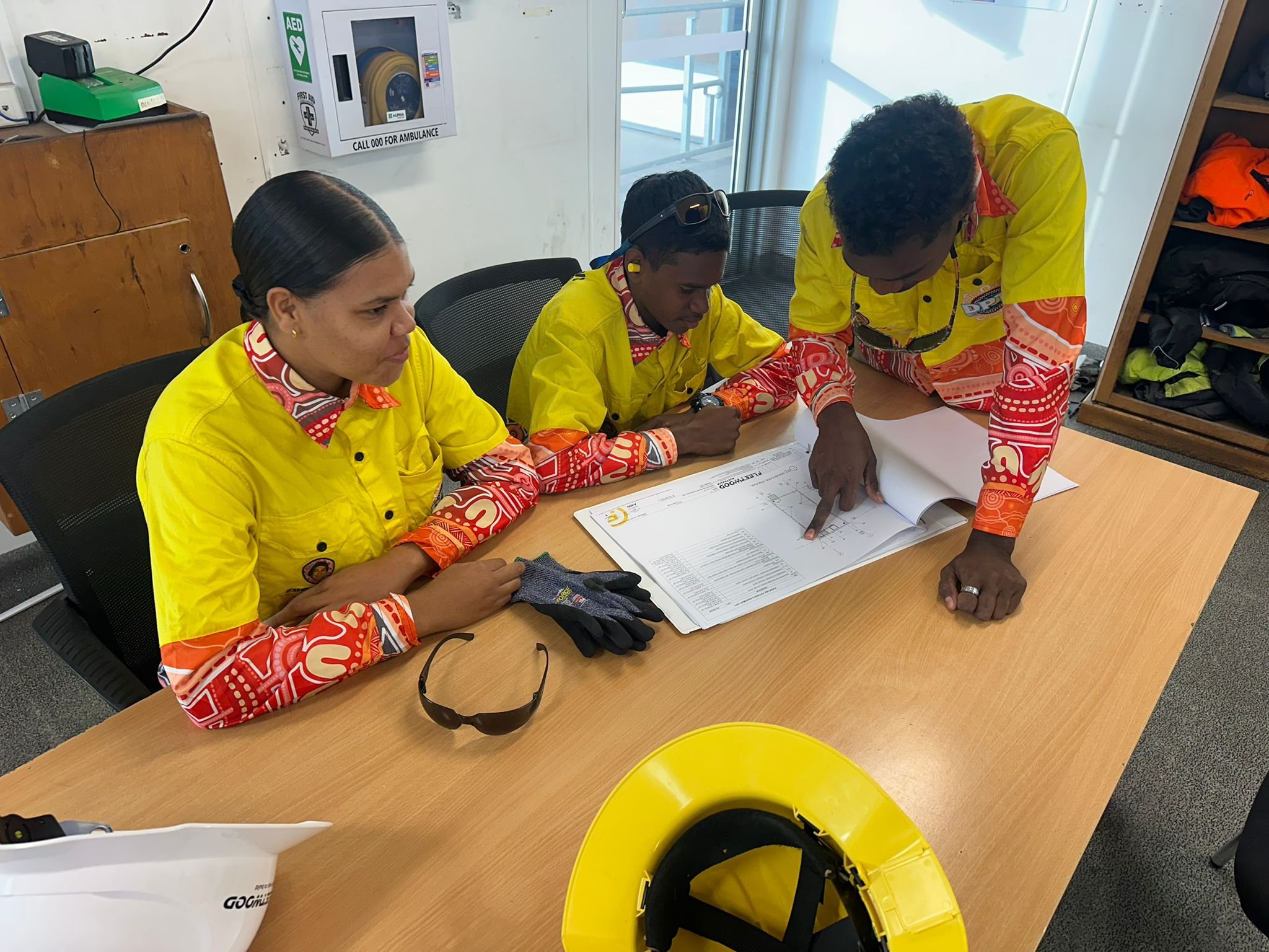 Three individuals wearing yellow safety uniforms gathered around a table reviewing documents. One person is pointing at the paper, and safety gear like gloves and a helmet are on the table. An AED cabinet and other safety equipment are visible on the wall.