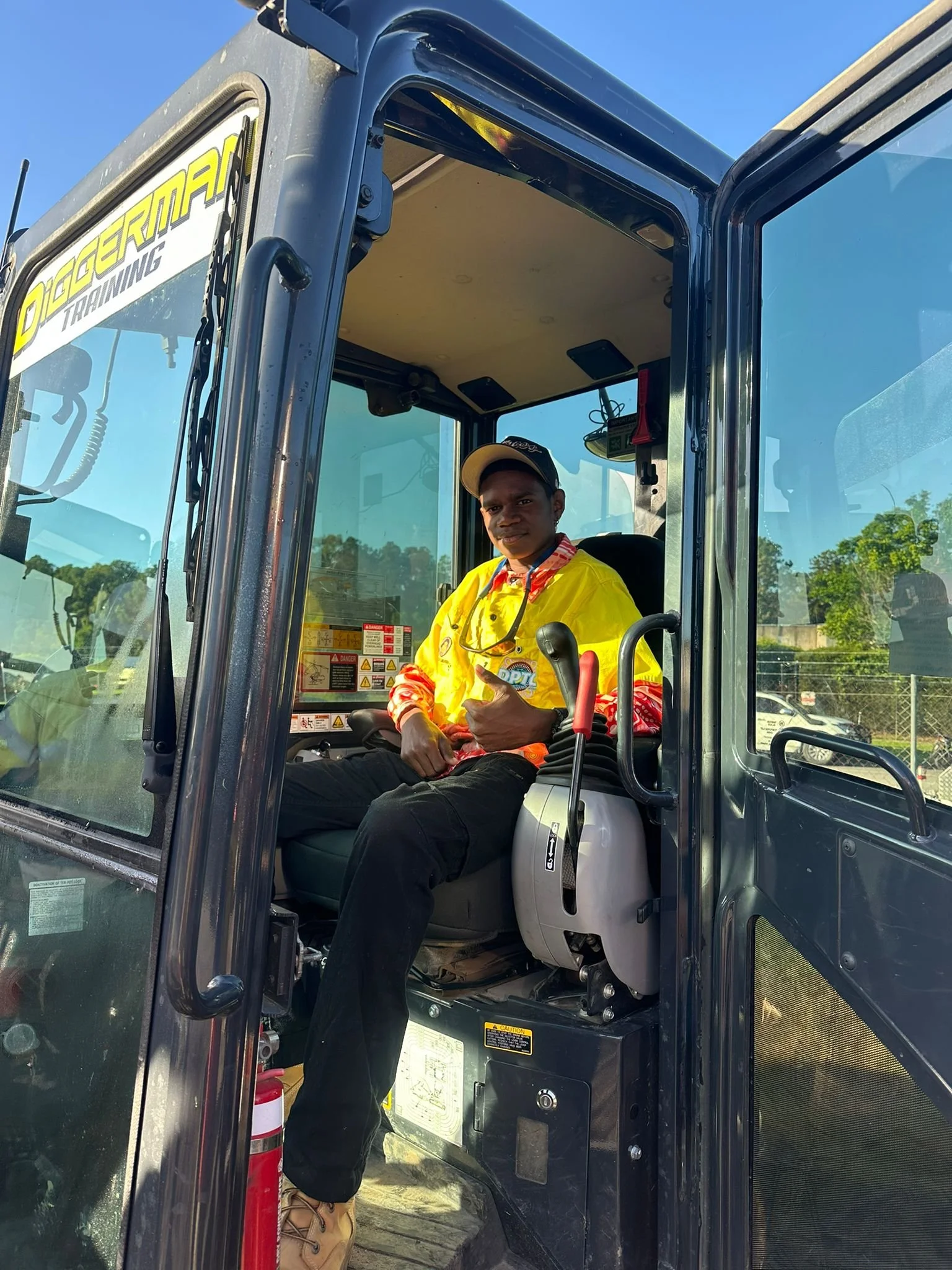 A person sitting in the driver's seat of a large construction vehicle, giving a thumbs-up gesture. The person is wearing a yellow high-visibility jacket, a black cap, and tan work boots. The image is taken from outside the vehicle with a clear day and trees in the background.