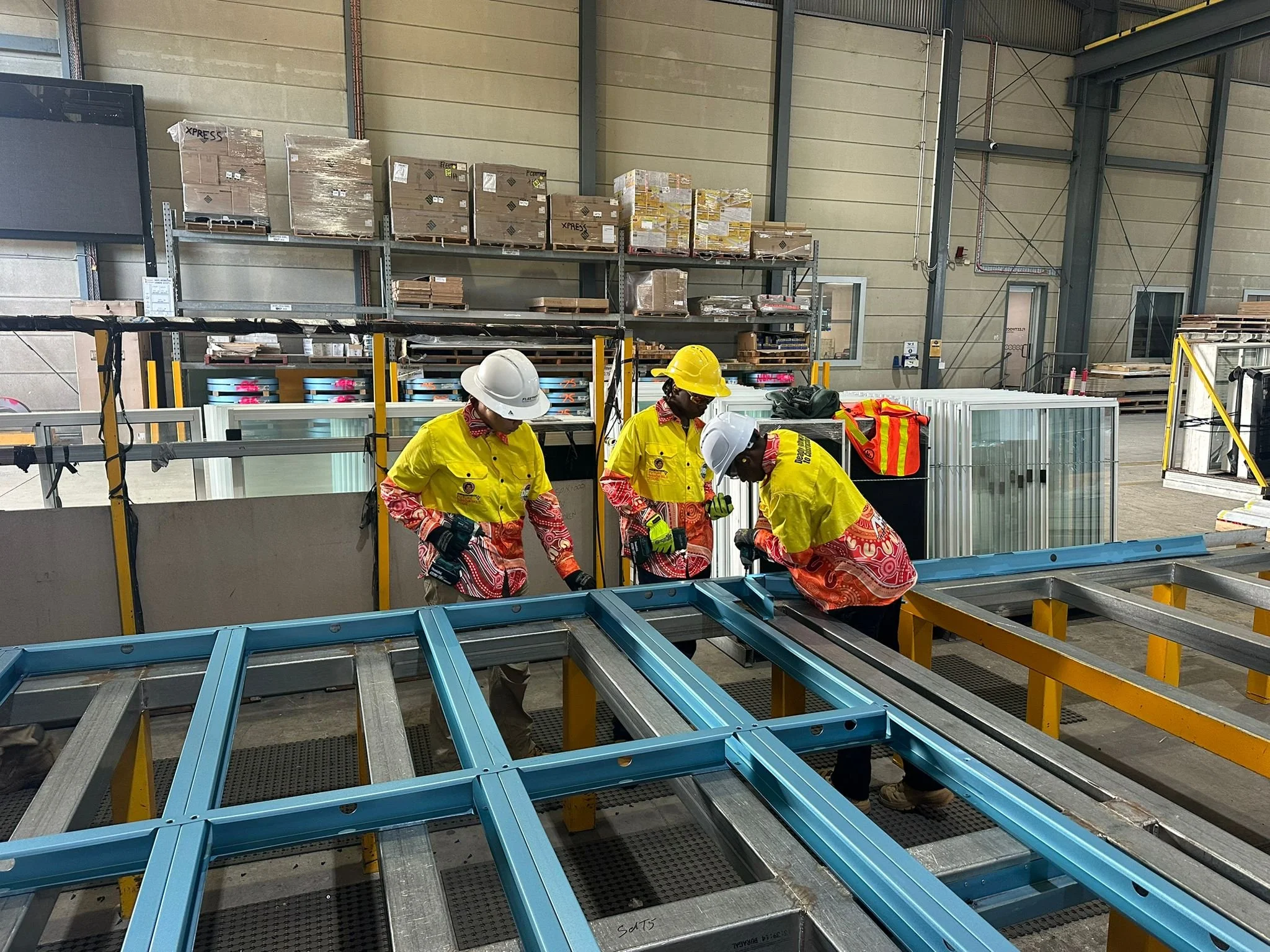 Workers wearing yellow safety gear and white helmets are assembling metal frames on a manufacturing line inside a warehouse. Shelves with boxes and pallets are visible in the background.