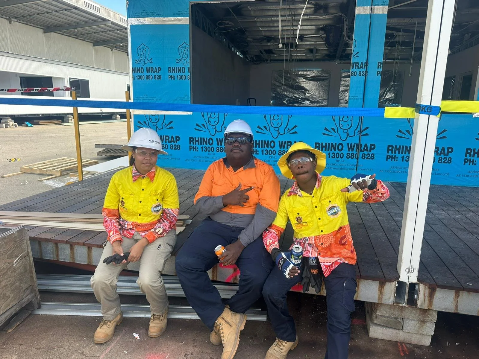Three construction workers sitting on a platform at a construction site, wearing safety helmets and gloves, with a partially built structure and blue Franken Wrap in the background.
