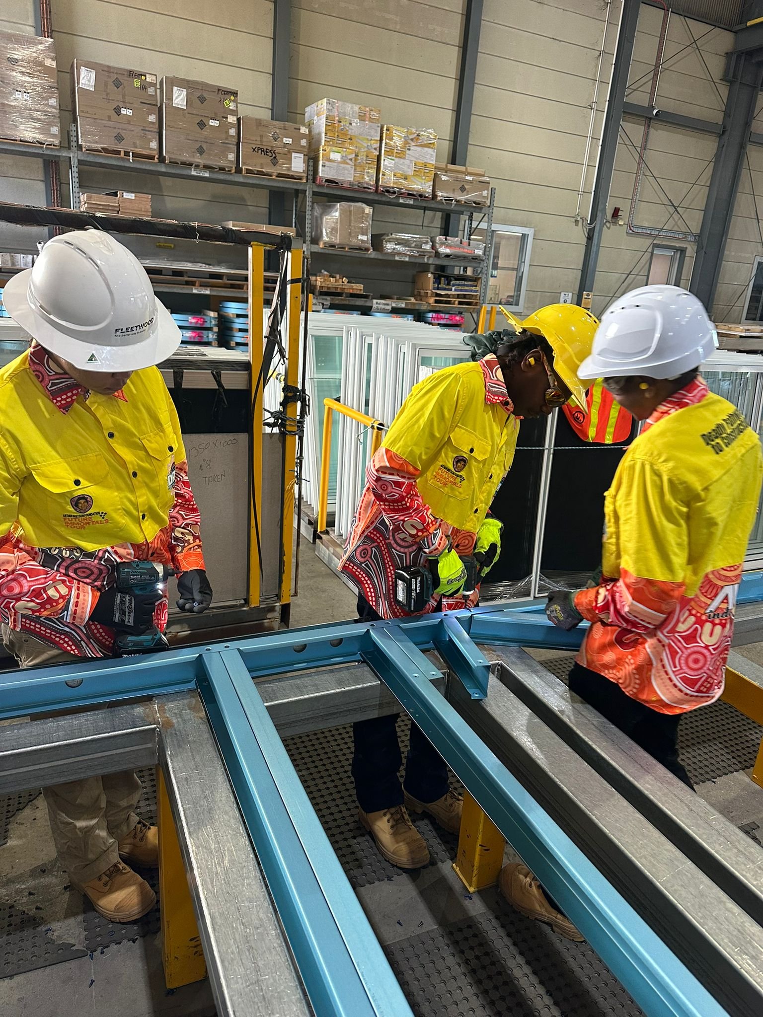 Workers in safety helmets and yellow shirts working with glass panels in a warehouse.