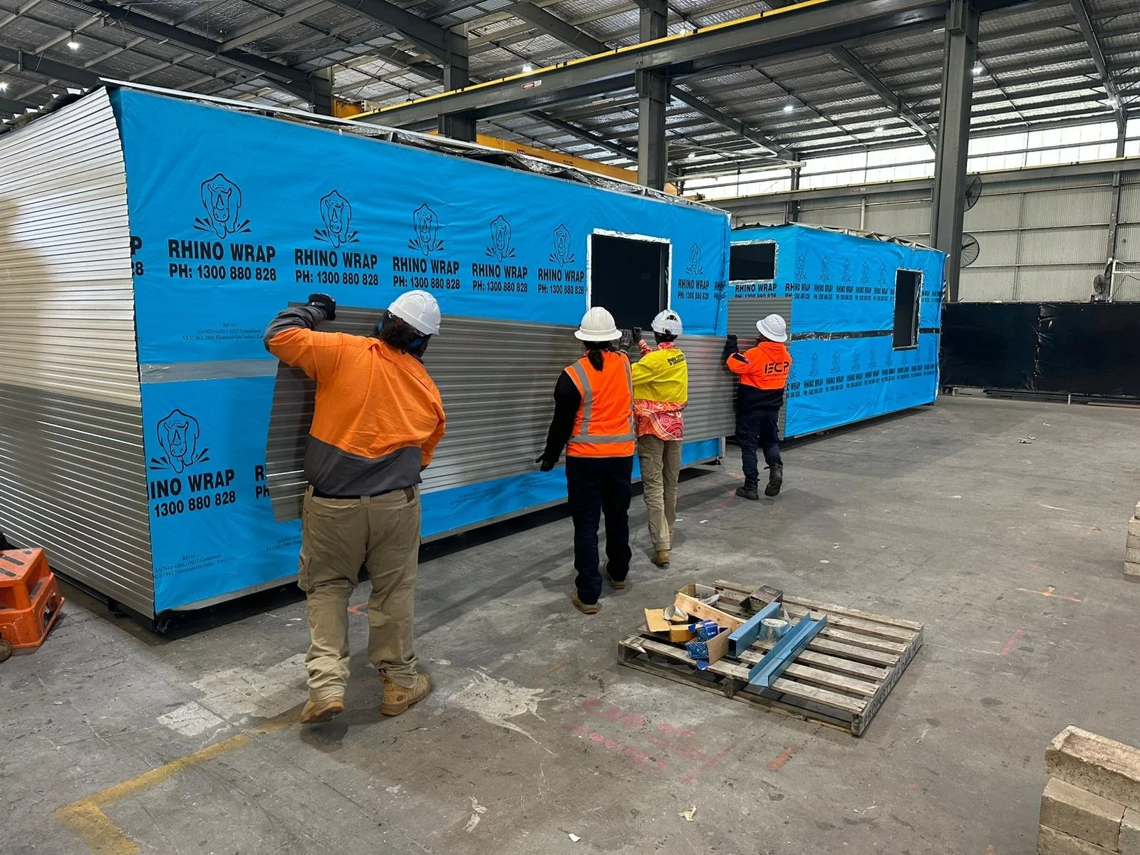 Construction workers wearing safety helmets and reflective vests installing metal panels on a large prefabricated structure inside an industrial warehouse.