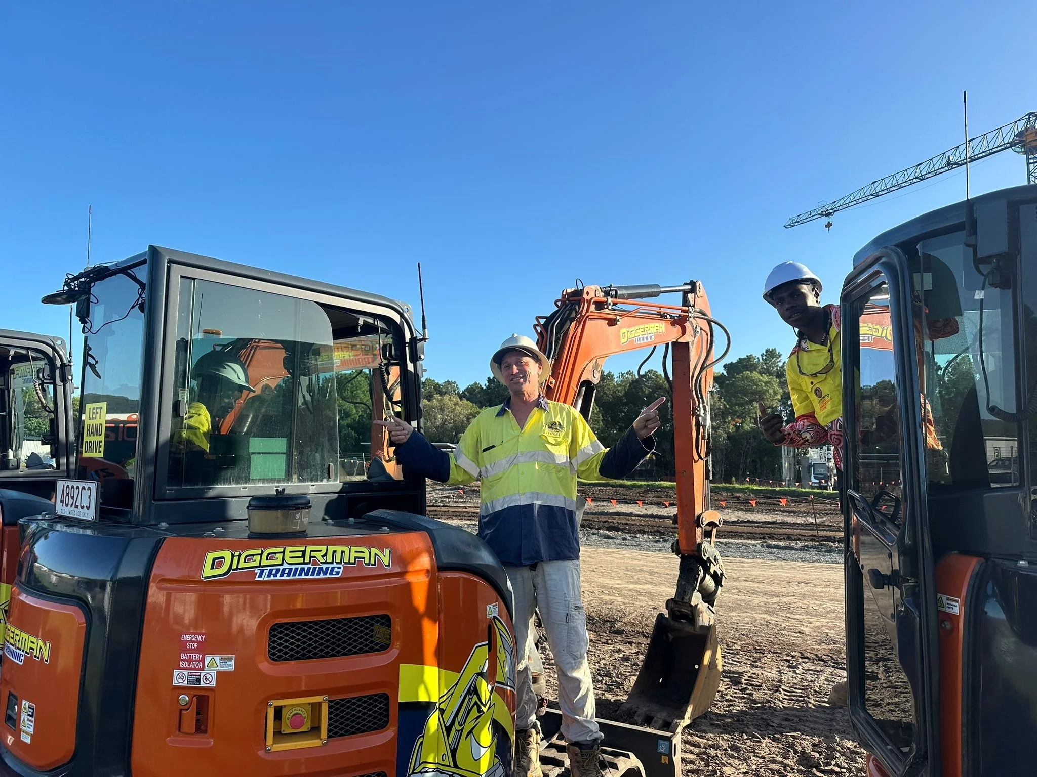 Two construction workers, one in a neon yellow safety jacket and the other in a yellow long-sleeve shirt, are standing next to orange digging equipment at an outdoor construction site. The worker in the safety jacket is smiling and pointing, while the other is holding a mobile phone. There are earth-moving machines and a crane visible in the background.