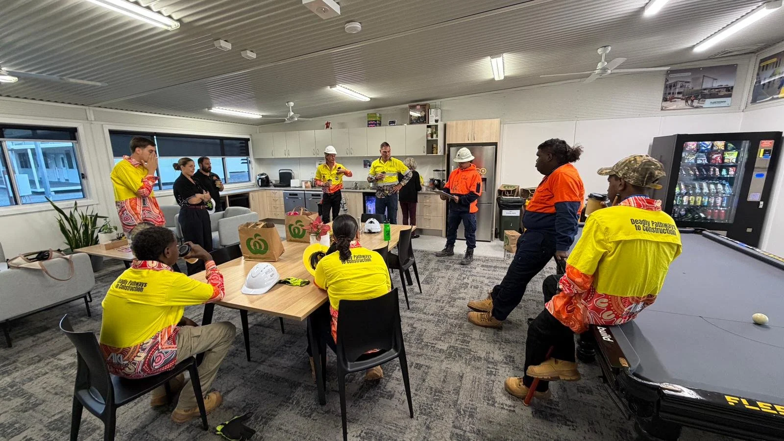 Workers in yellow and orange shirts gathered in a break room with a table, vending machines, and a pool table, having a meeting or break.