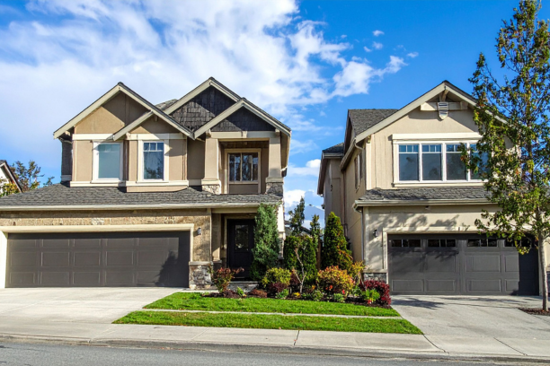Two modern suburban houses with front lawns and garages under a partly cloudy sky.