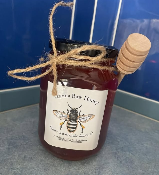 Jar of raw honey with a bee illustration on the label, a wooden honey dipper attached to the lid, and a twine bow around the neck, placed on a countertop.