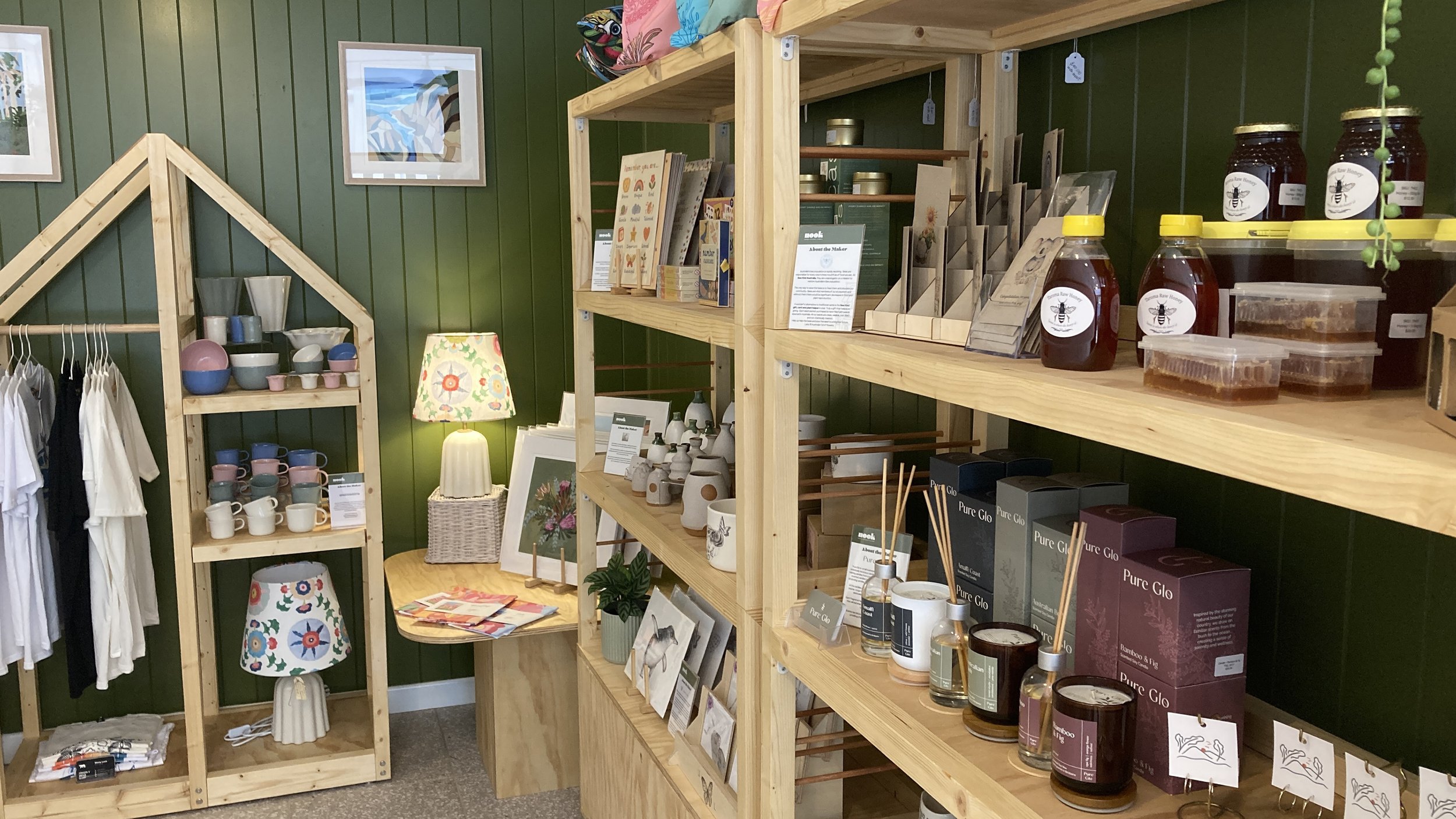 Retail display with colorful ceramic cups, mugs, and table lamps, along with skincare products, candles, and decorative items on wooden shelves against a green wall.
