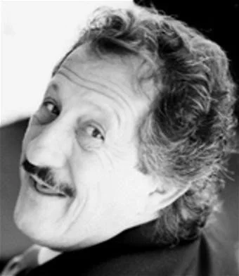 Black and white photo of a smiling man with wavy hair and mustache, looking back over his shoulder.