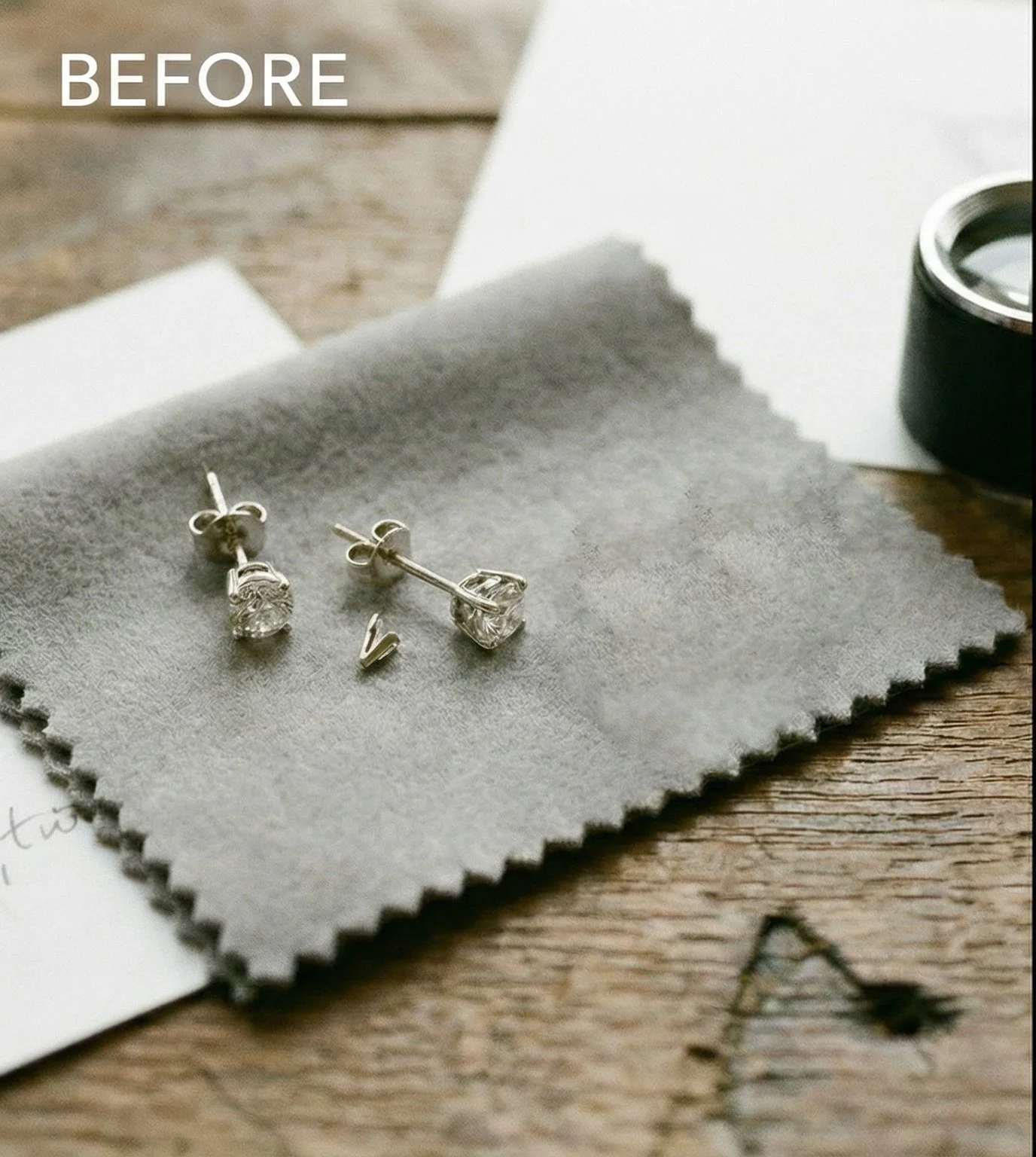 A pair of silver earrings with clear gemstones resting on a gray fabric, with a wooden surface and a roll of black tape nearby.