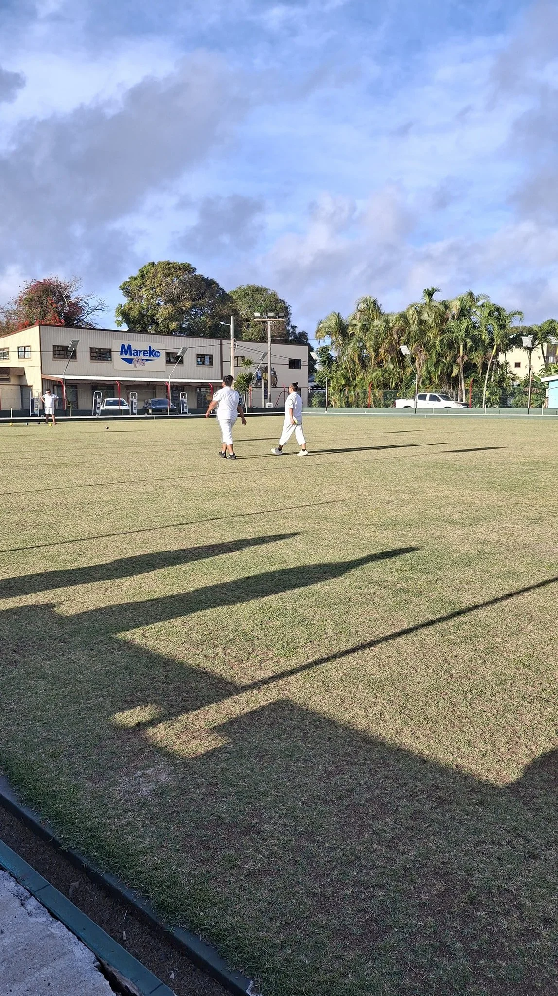 People dressed in white playing lawn bowls on a well-maintained grass field with a building labeled 'Mareko' in the background, bordered by trees and parked cars under a partly cloudy sky.