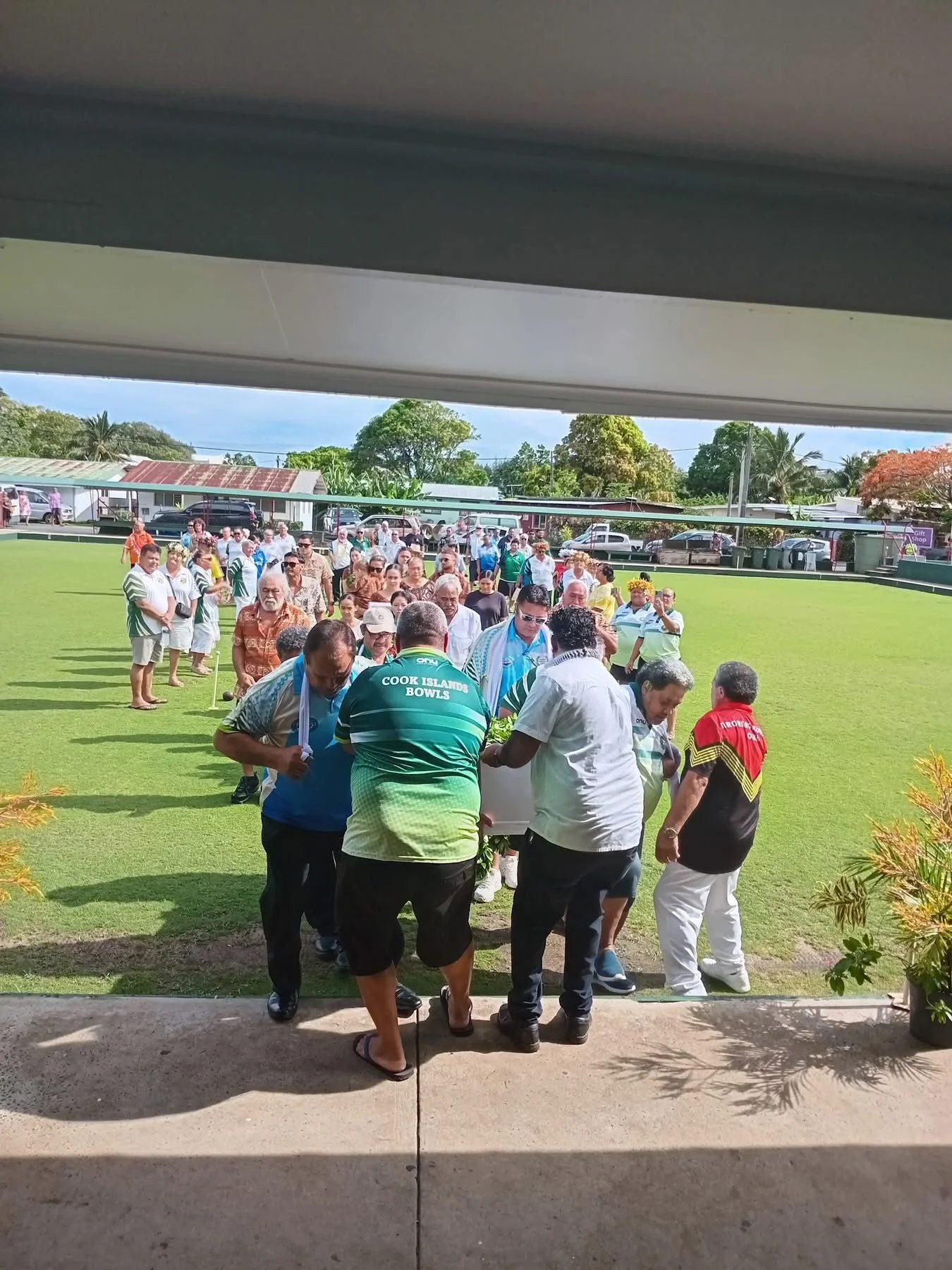 A group of people gathered outside on a grassy field for a ceremonial event, with some individuals carrying a floral arrangement.