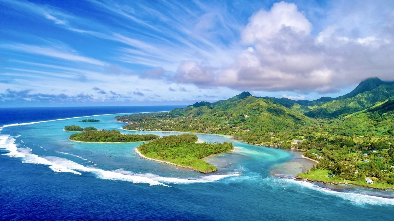 Aerial view of a tropical landscape with lush green islands, clear blue ocean, and mountains in the background on a partly cloudy day.