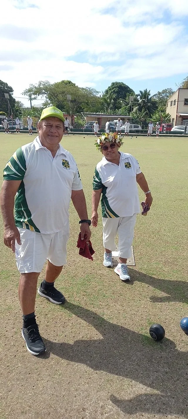 Two older adults playing lawn bowls outside on a sunny day, wearing white uniforms with green and yellow accents, one with a flower crown and sunglasses, others in the background, and trees and buildings around.