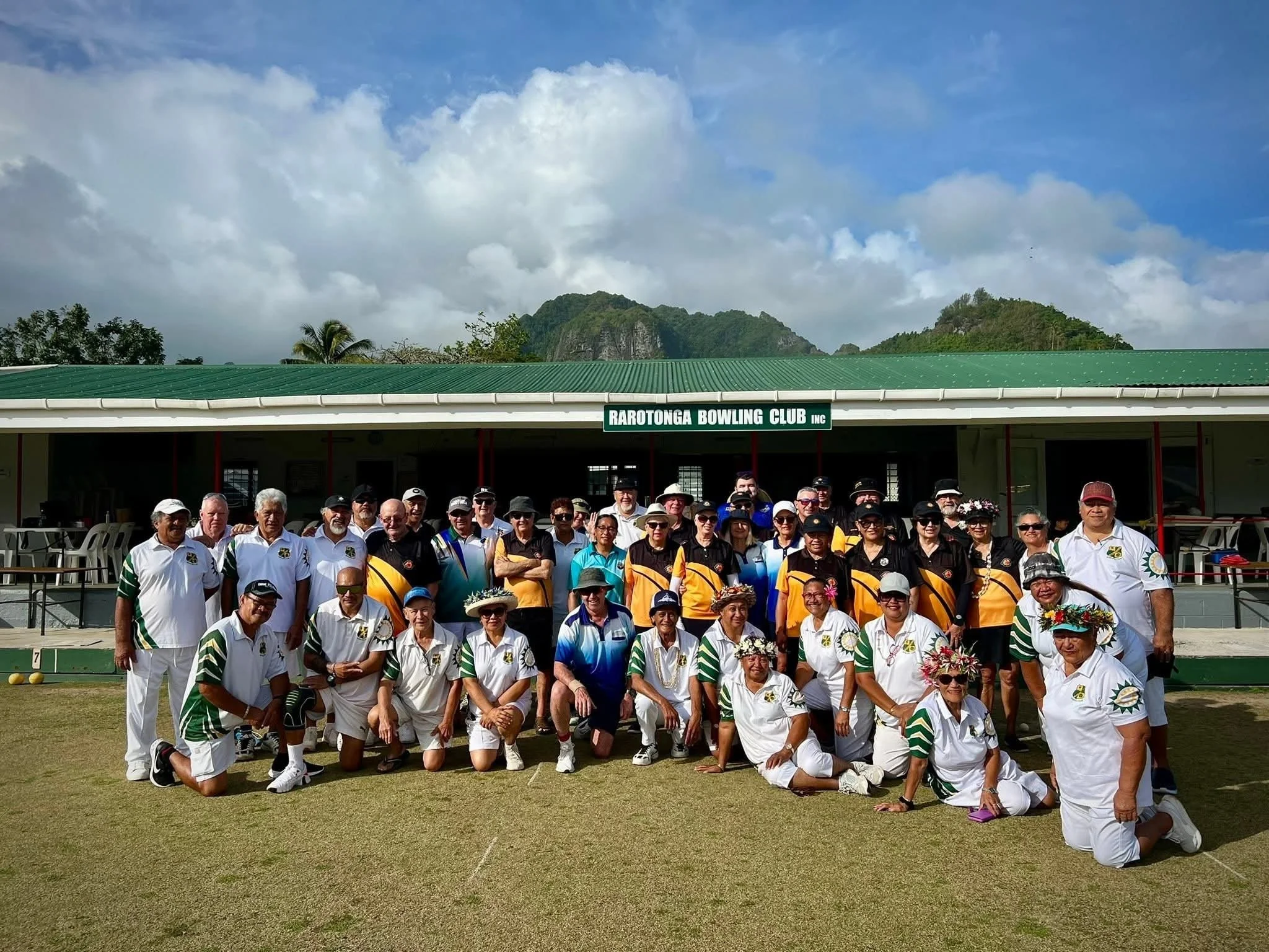 A group of people gathered at the Rarotonga Bowling Club in front of a building with a sign, with mountains and a cloudy sky in the background.