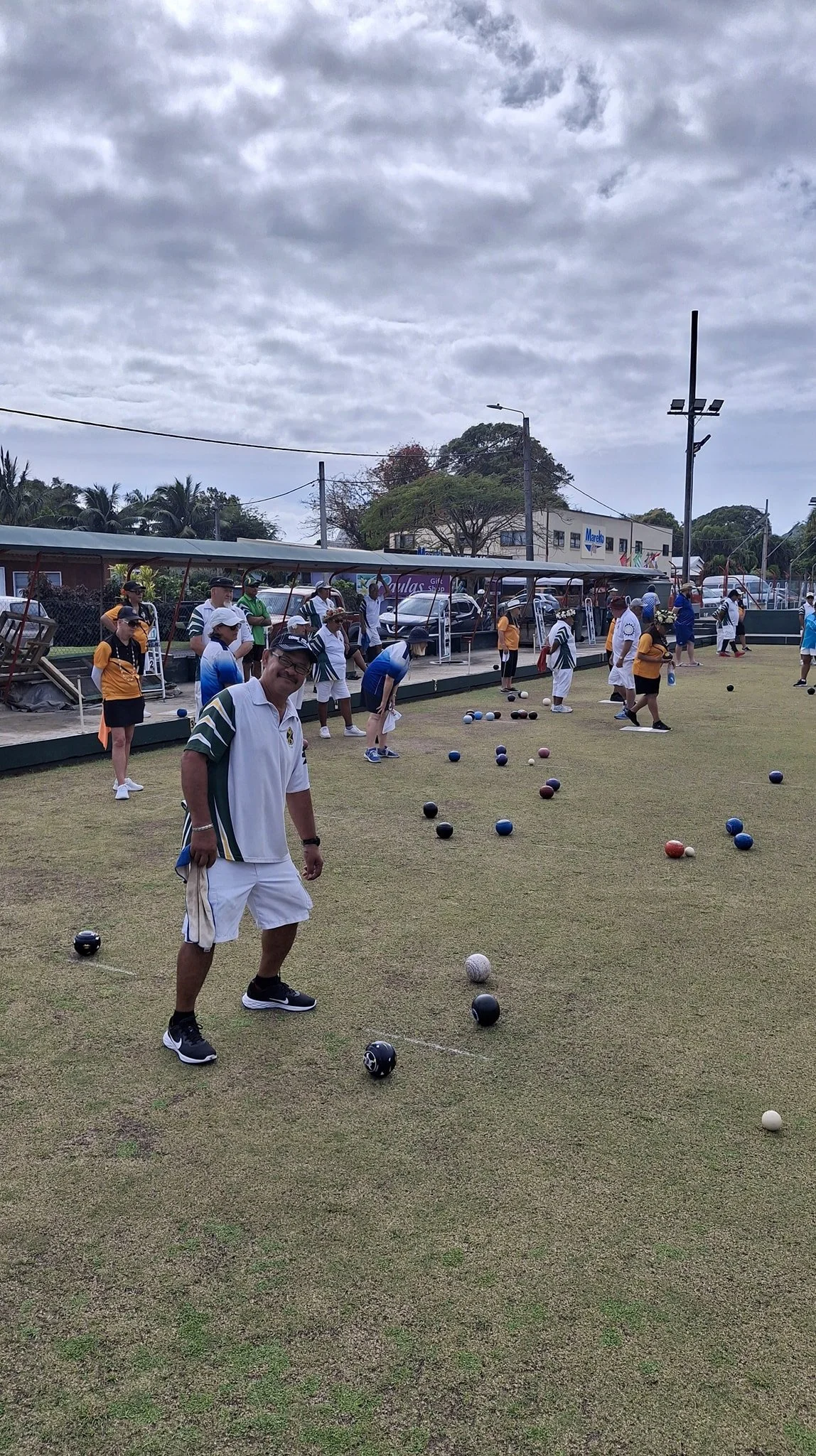 People playing lawn bowls on a grassy field under cloudy sky, with some spectators and parked cars in the background.