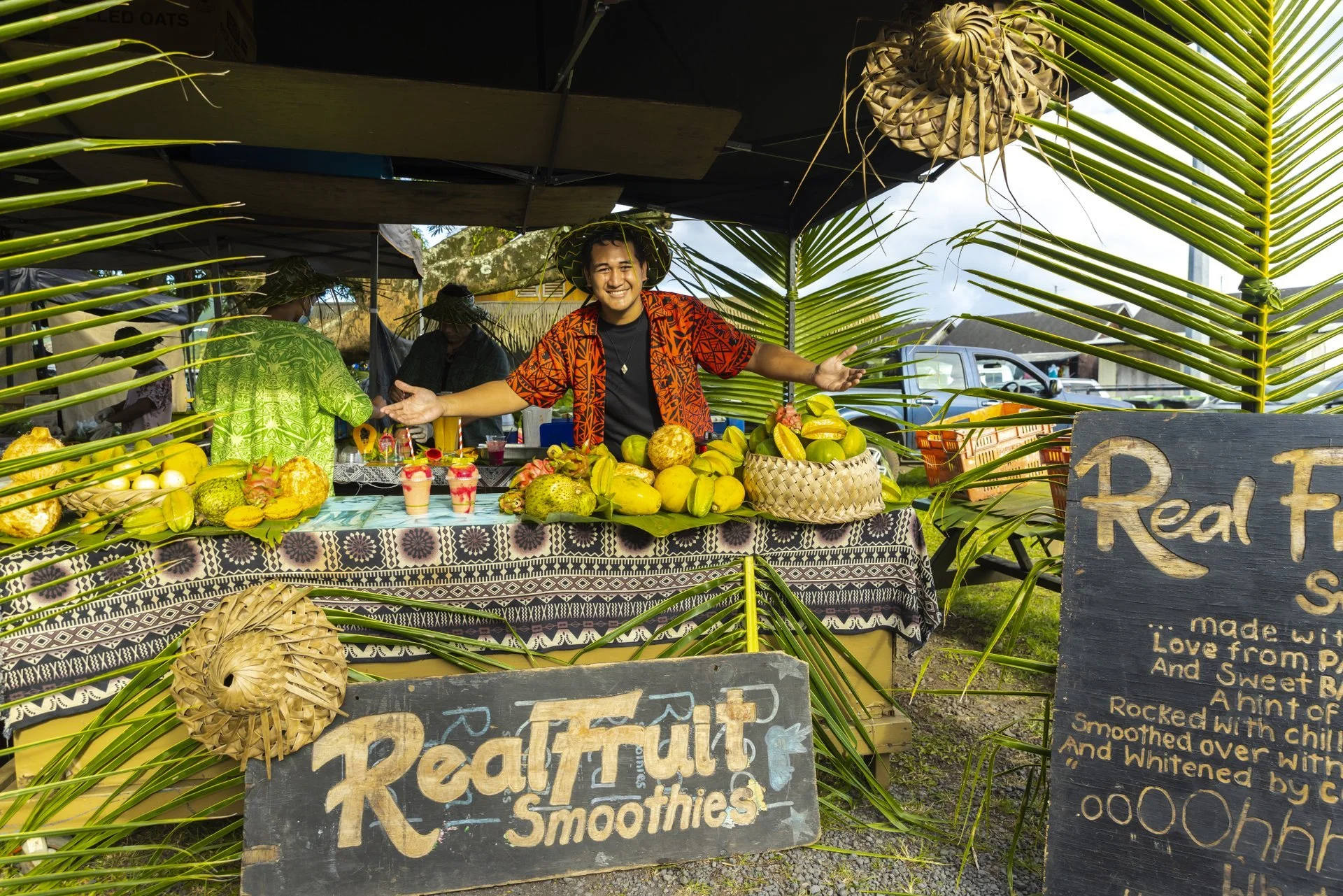 A smiling man at a fruit smoothie stall decorated with tropical leaves and fruits like starfruit, guava, and papaya, with a handwritten sign advertising real fruit smoothies.